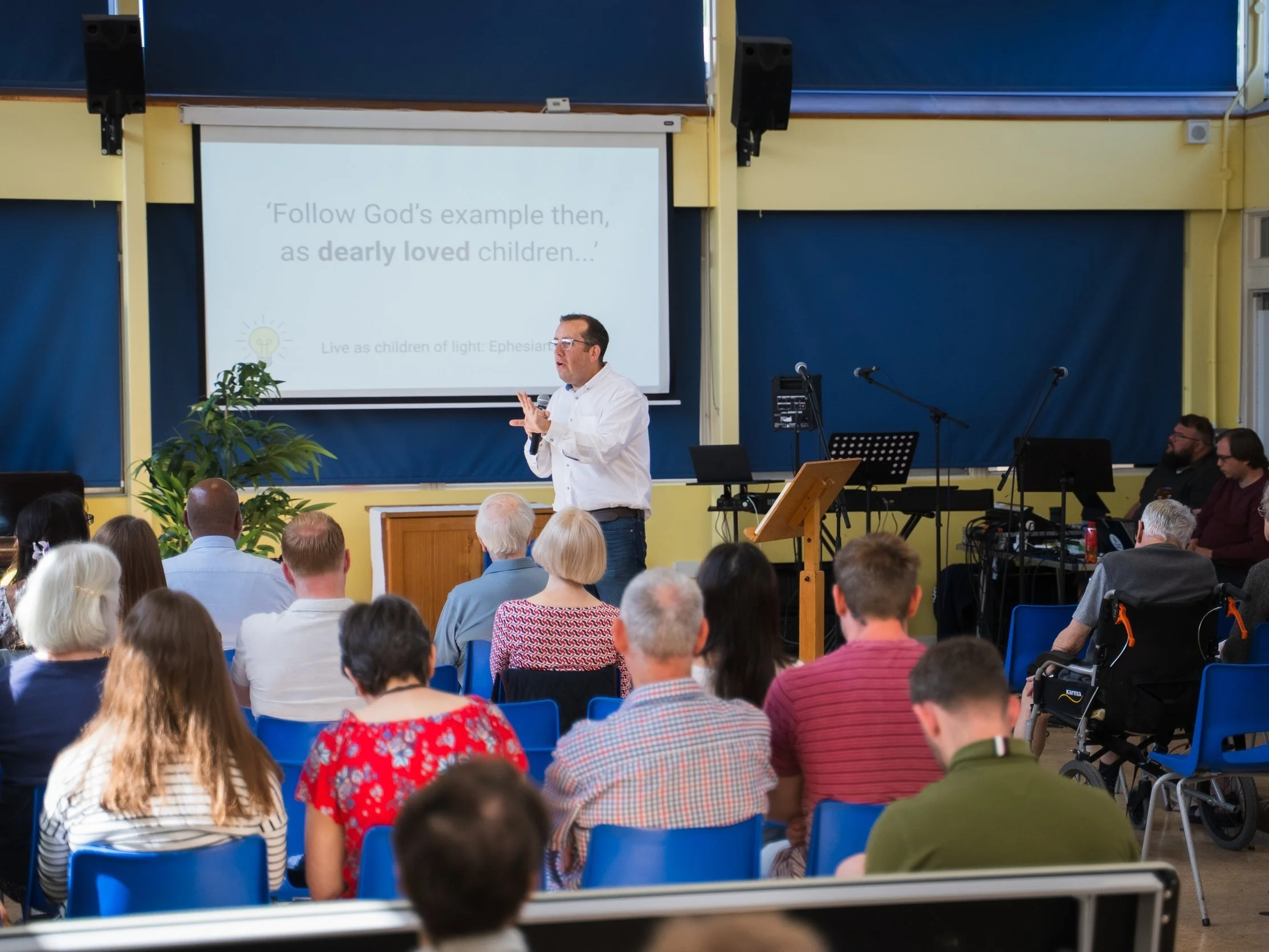 A man giving a presentation in a classroom or conference room filled with seated audience members. A large screen behind him displays a slide with text.
