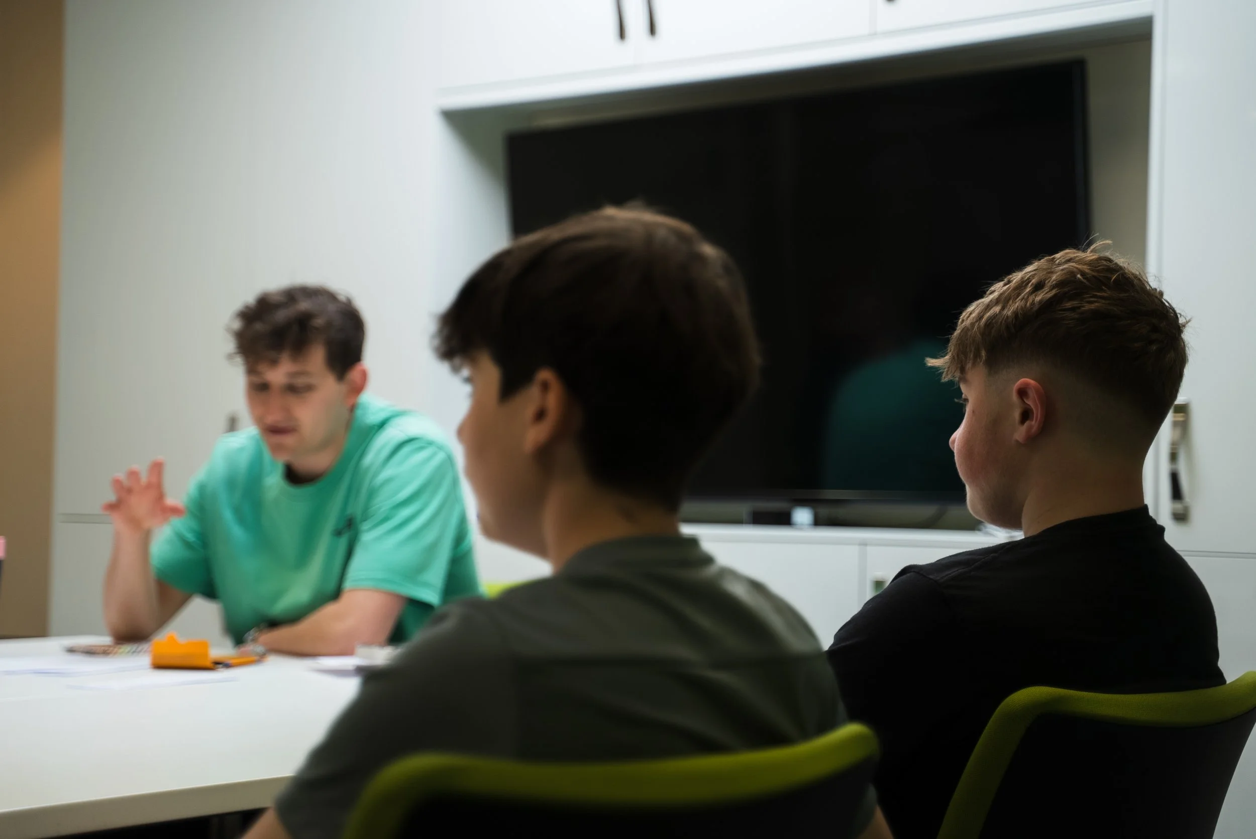Three young men sit at a table in a room with a television in the background, engaged in discussion. One man in a green shirt is speaking or gesturing, while the other two listen.