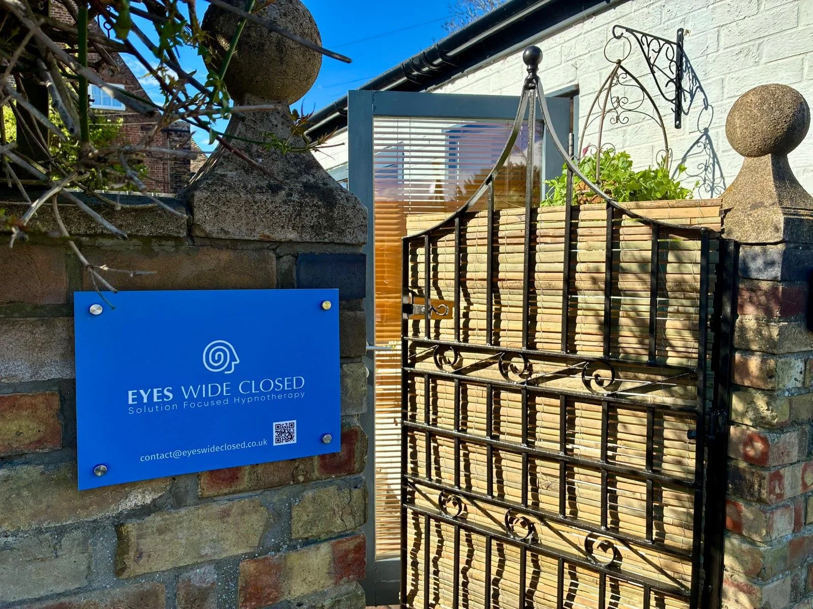 A brick wall with a blue sign that reads 'Eyes Wide Closed, Solution Focused Hypnotherapy,' with an email and a QR code. There is a black wrought iron gate with decorative scrollwork and a wooden privacy screen behind it, with sunlight casting shadows.