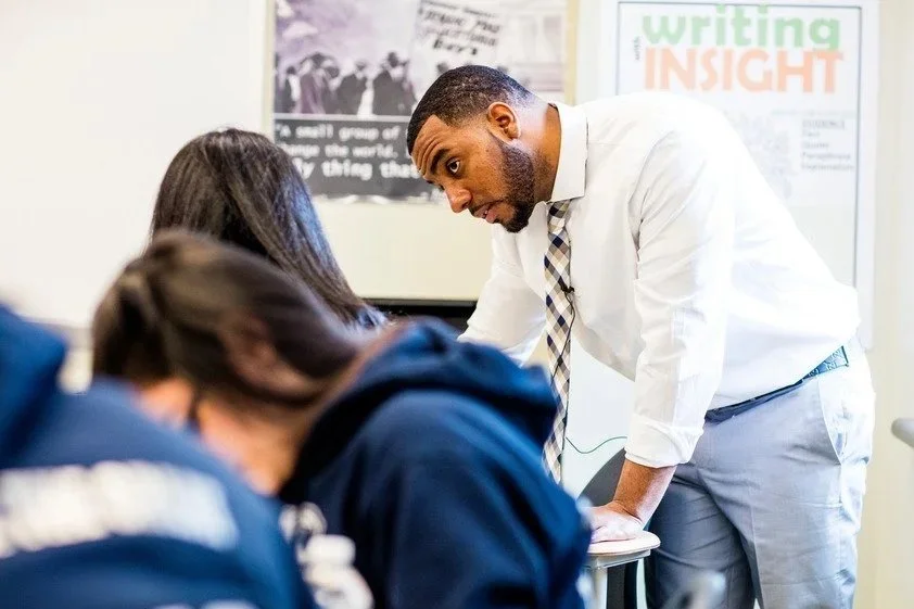 Two people in a meeting room having a discussion. The woman is wearing a dark blue polka dot shirt and the man is in a light blue shirt. They are seated at a table with papers and a mug visible.