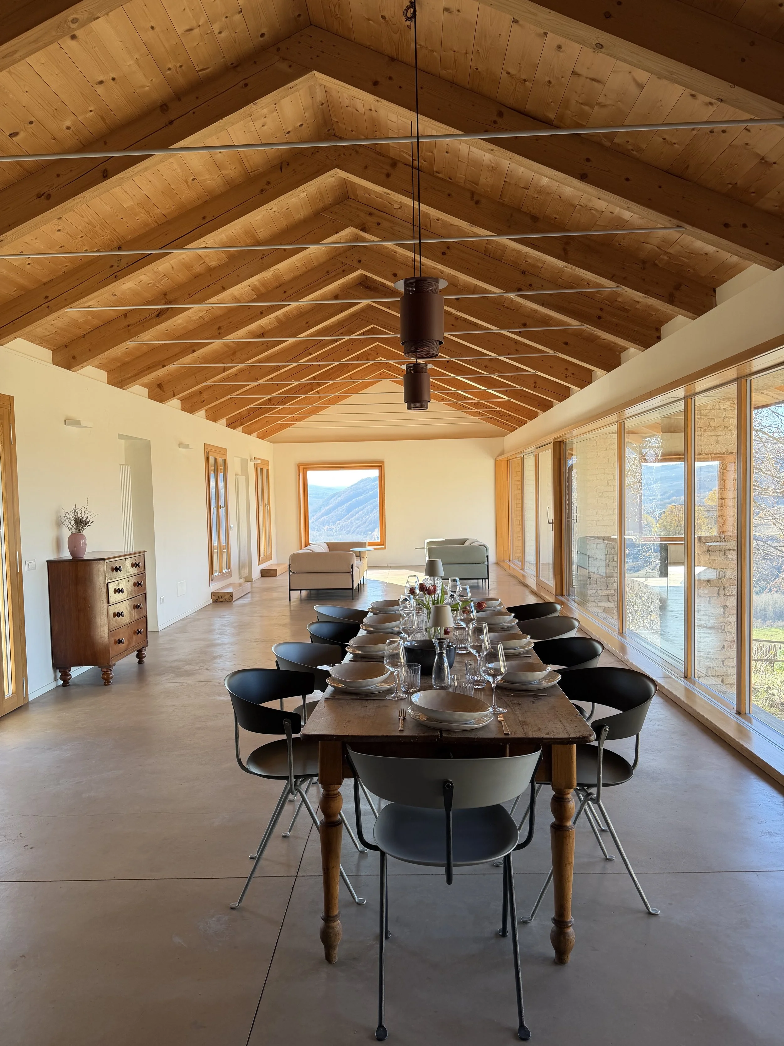 Sala da pranzo con tavola apparecchiata, sedie nere, soffitto in legno a vista, grandi finestre con vista sulle montagne.