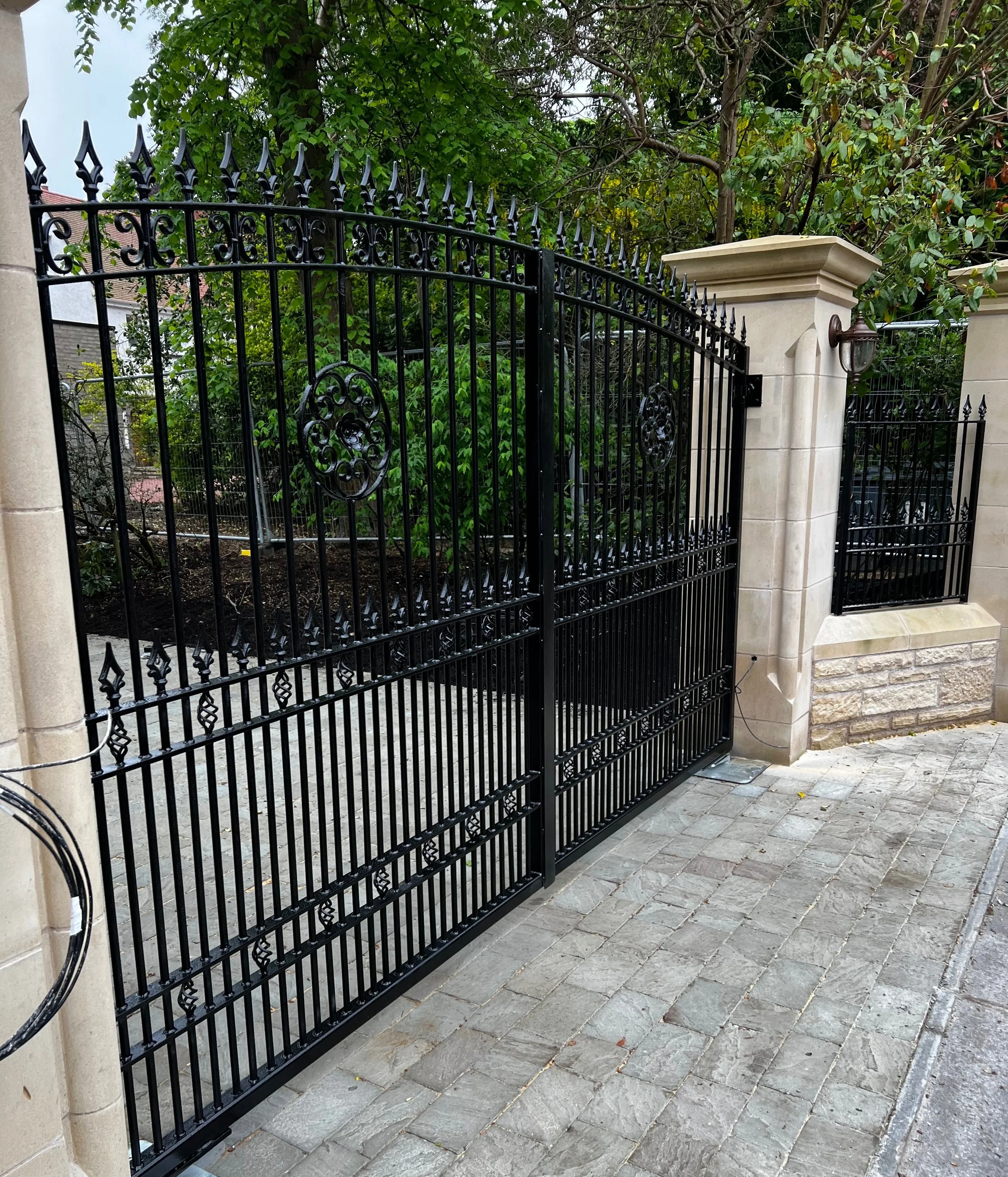 Black wrought iron gate with decorative scrolls and pointed finials, supported by beige stone columns, leading to a garden area with green trees, and a small brick-paved sidewalk in the foreground.