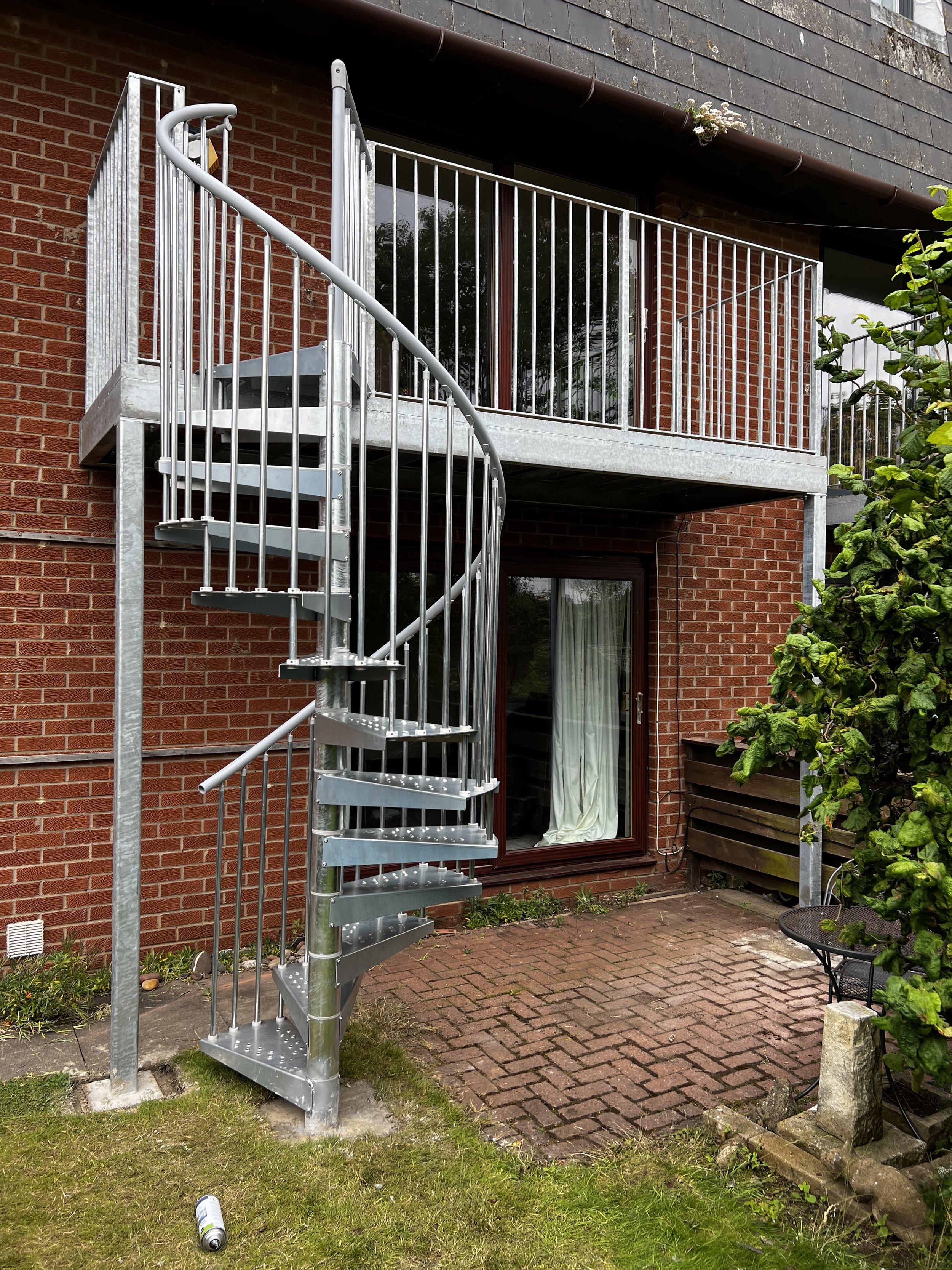 A metal spiral staircase with safety railings in a backyard next to a brick house with a sliding glass door. There are some plants and a round metal table with chairs to the right, and a grass and brick patio area.
