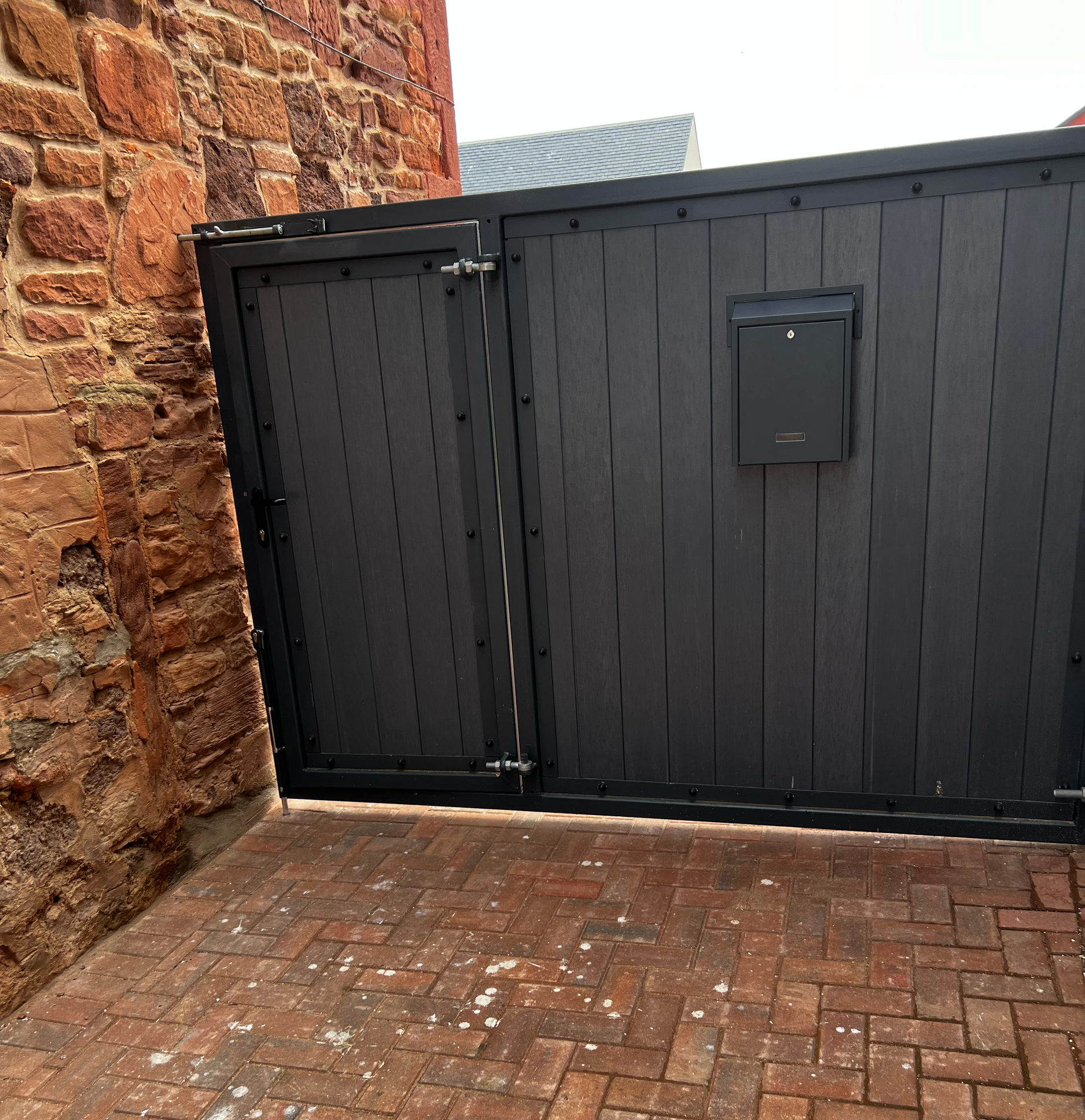 A black wooden gate with metal hinges and latch, partially open, situated in an outdoor brick courtyard next to a red brick stone wall.
