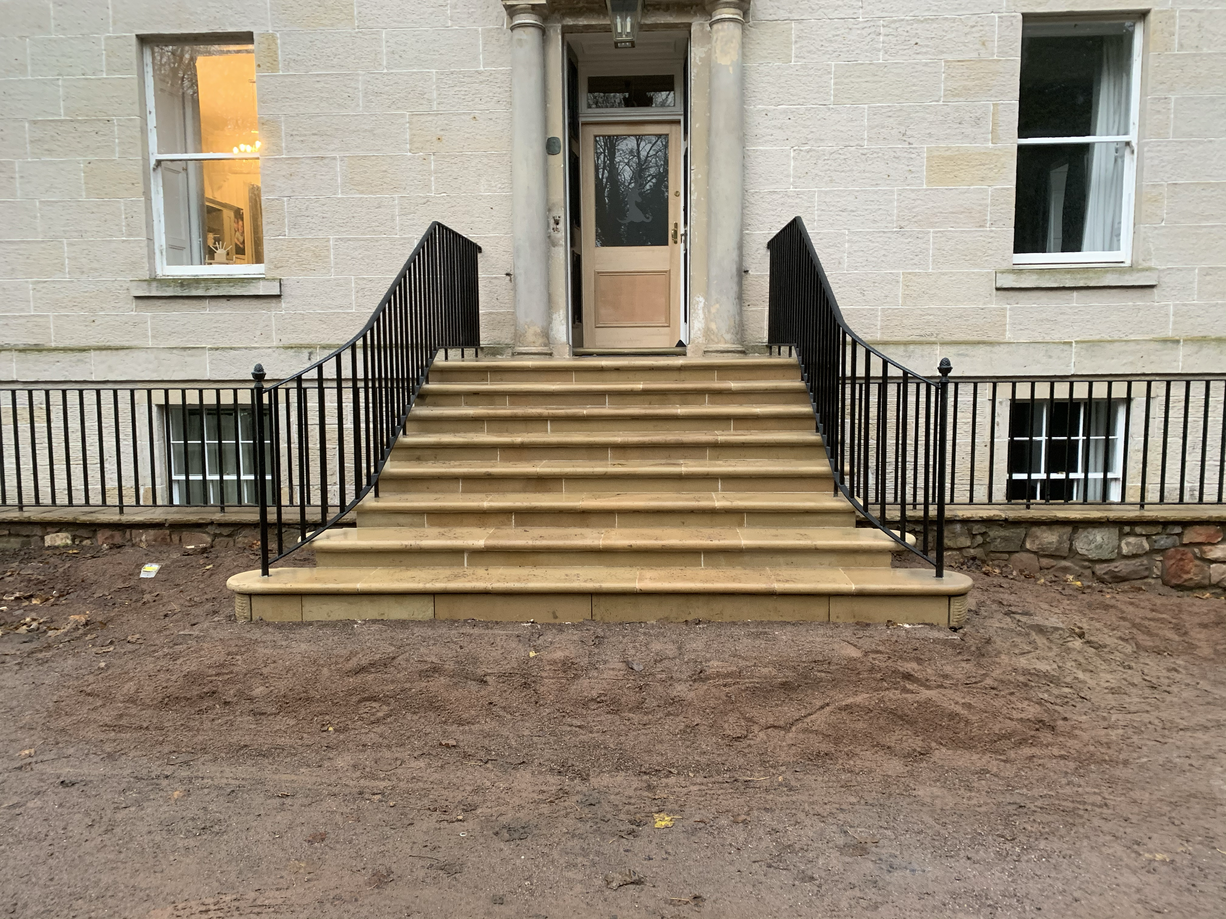 Front entrance of a stone building with a staircase, black metal railings, and a tan wooden door. Two windows are visible, one on each side of the door. The left window shows a warmly lit interior, and the right window shows part of a white curtain inside.