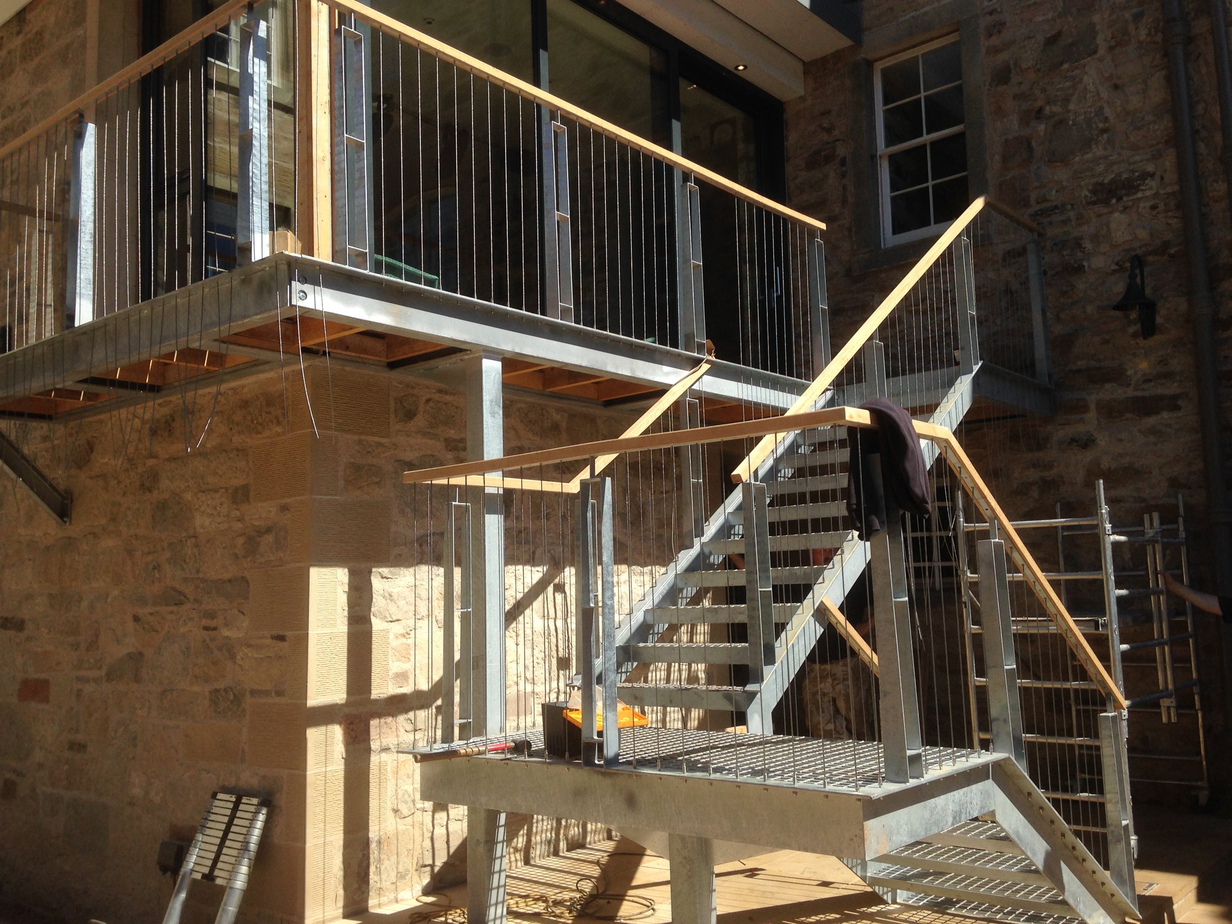 A metal stairway under construction with unfinished wooden handrails, located outside a stone building with a window.