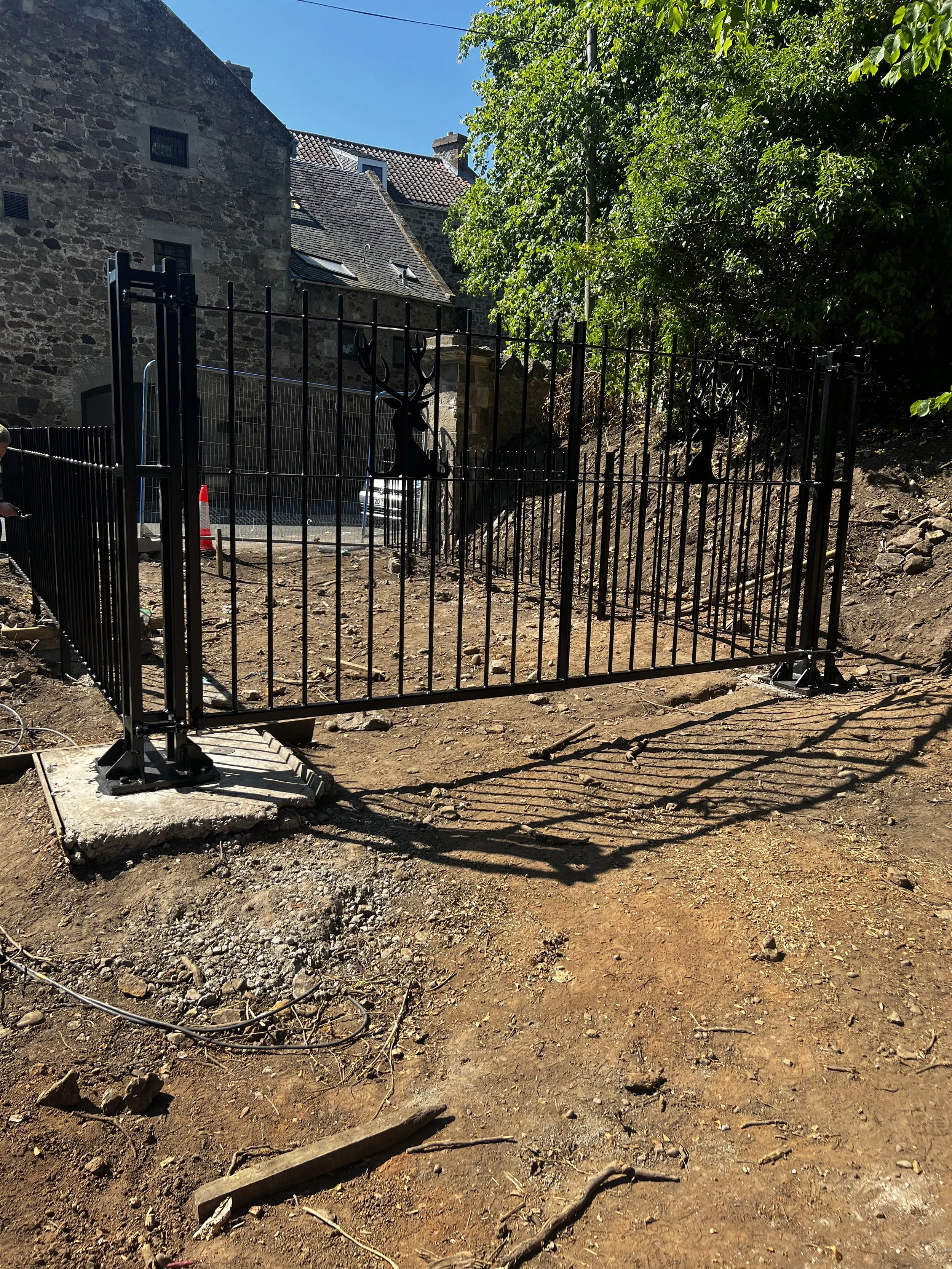 View of a construction site with a black metal fence, dirt ground, and a building with stone walls and a tiled roof in the background, under a clear blue sky.