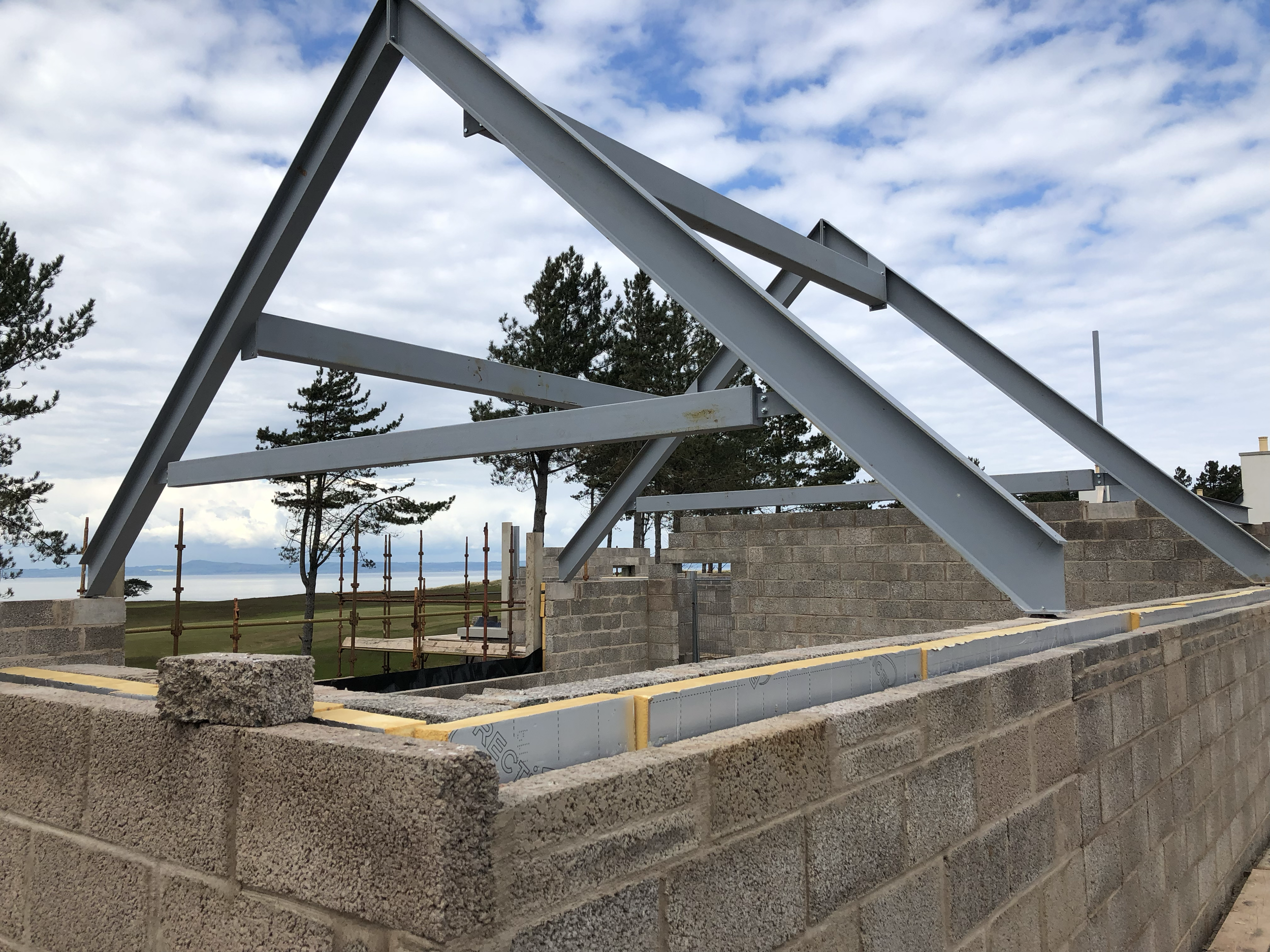 Construction site with brick wall framing and silver metal beams against a cloudy sky with trees and water in the background.