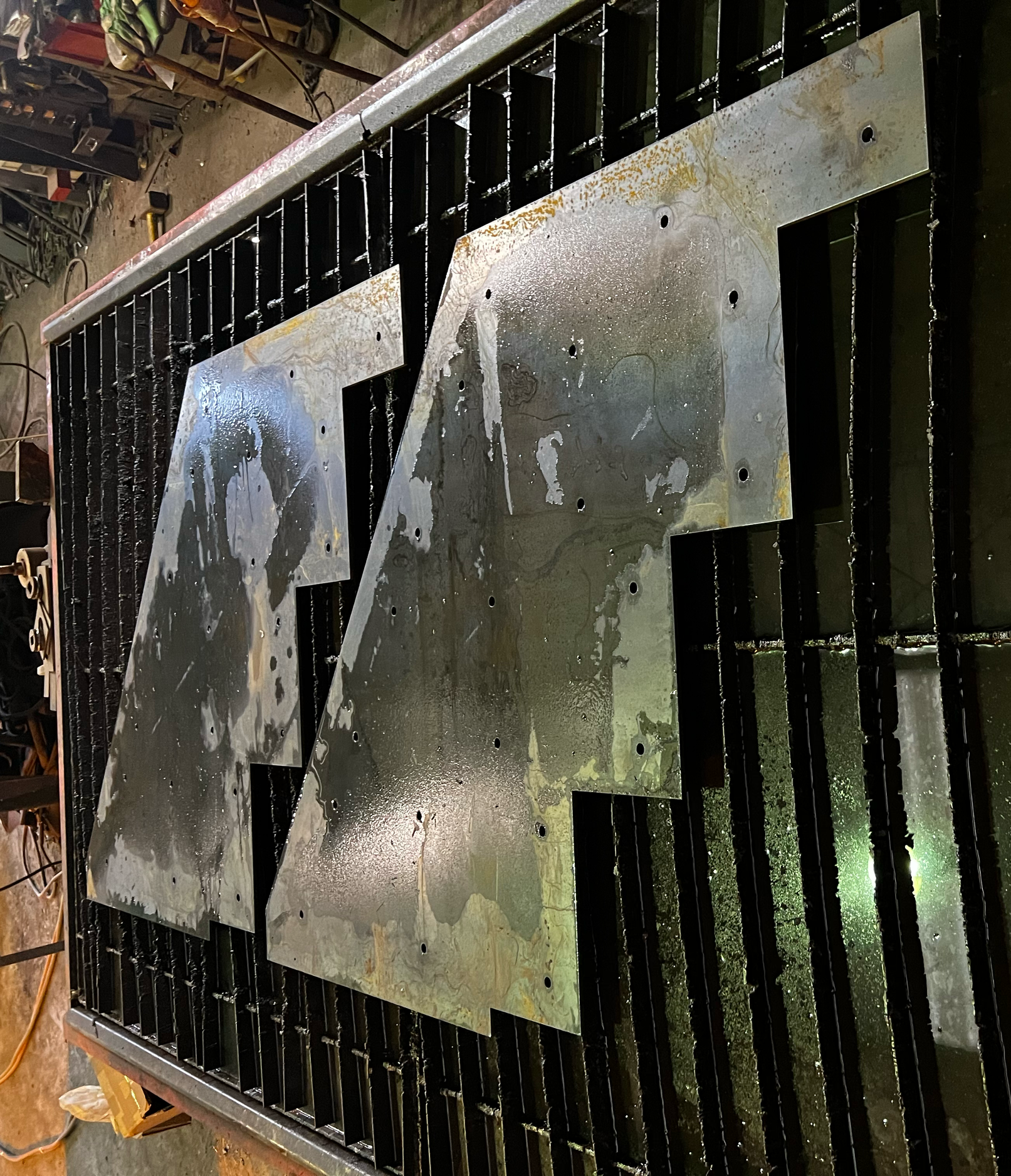 Metal sheets with drilled holes resting on a grid in a workshop, with tools and equipment visible in the background.