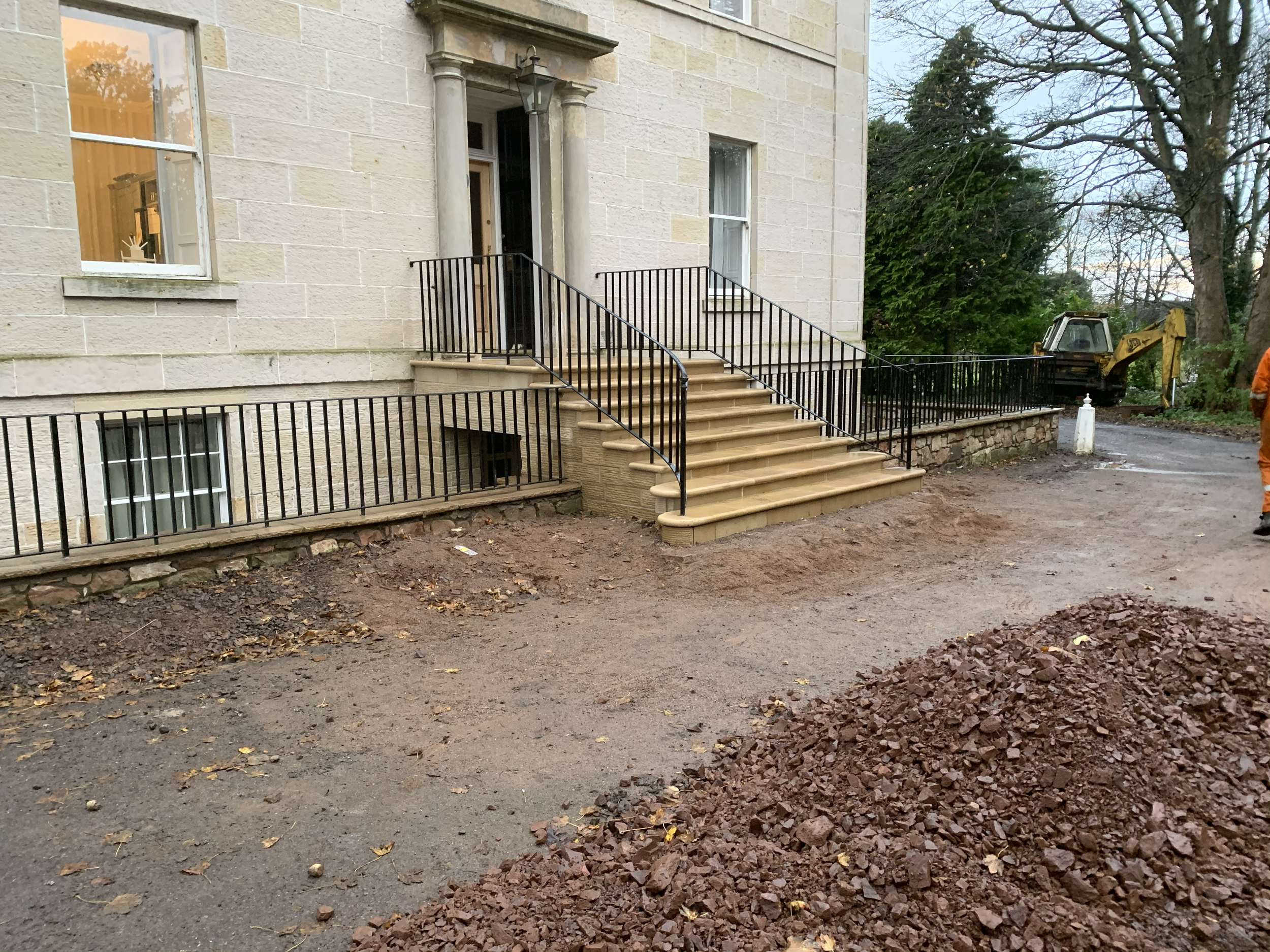 A house with a stone exterior, front steps with black metal railings, and a door with columns on either side. There are large trees surrounding the house, and construction equipment is on the right side.