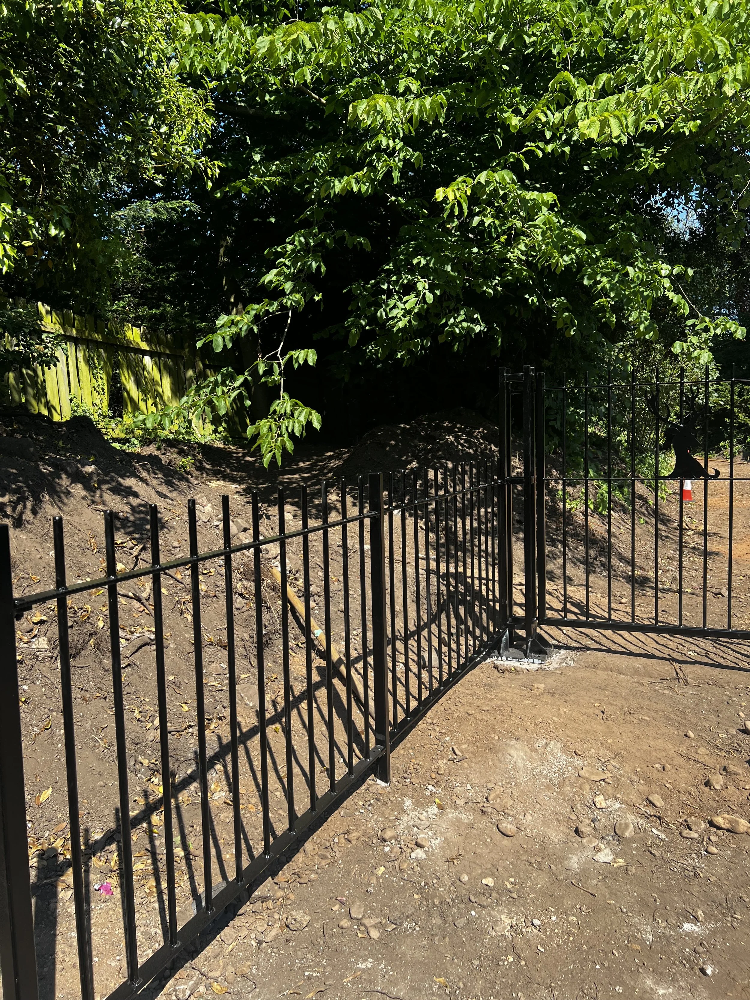 A black metal fence surrounds a dug-up patch of dirt and gravel. Behind the fence is a large, leafy green tree and a wooden fence to the left. The scene appears to be an outdoor area, possibly under construction or landscaping.