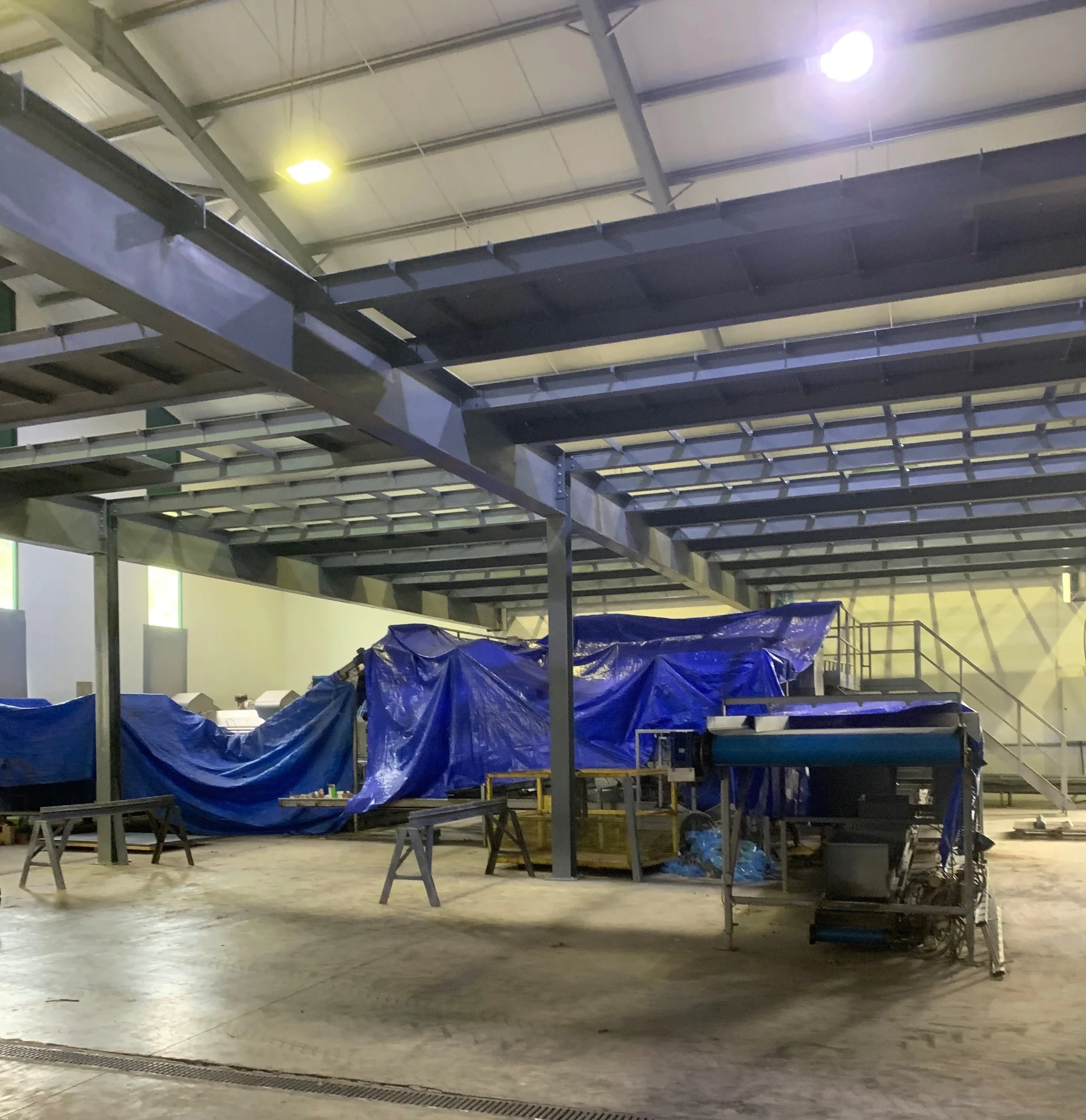 A large indoor space with a high ceiling, steel beams, and industrial lighting. There is a metal structure, possibly a storage or work platform, with blue tarps covering something underneath. The floor is concrete.