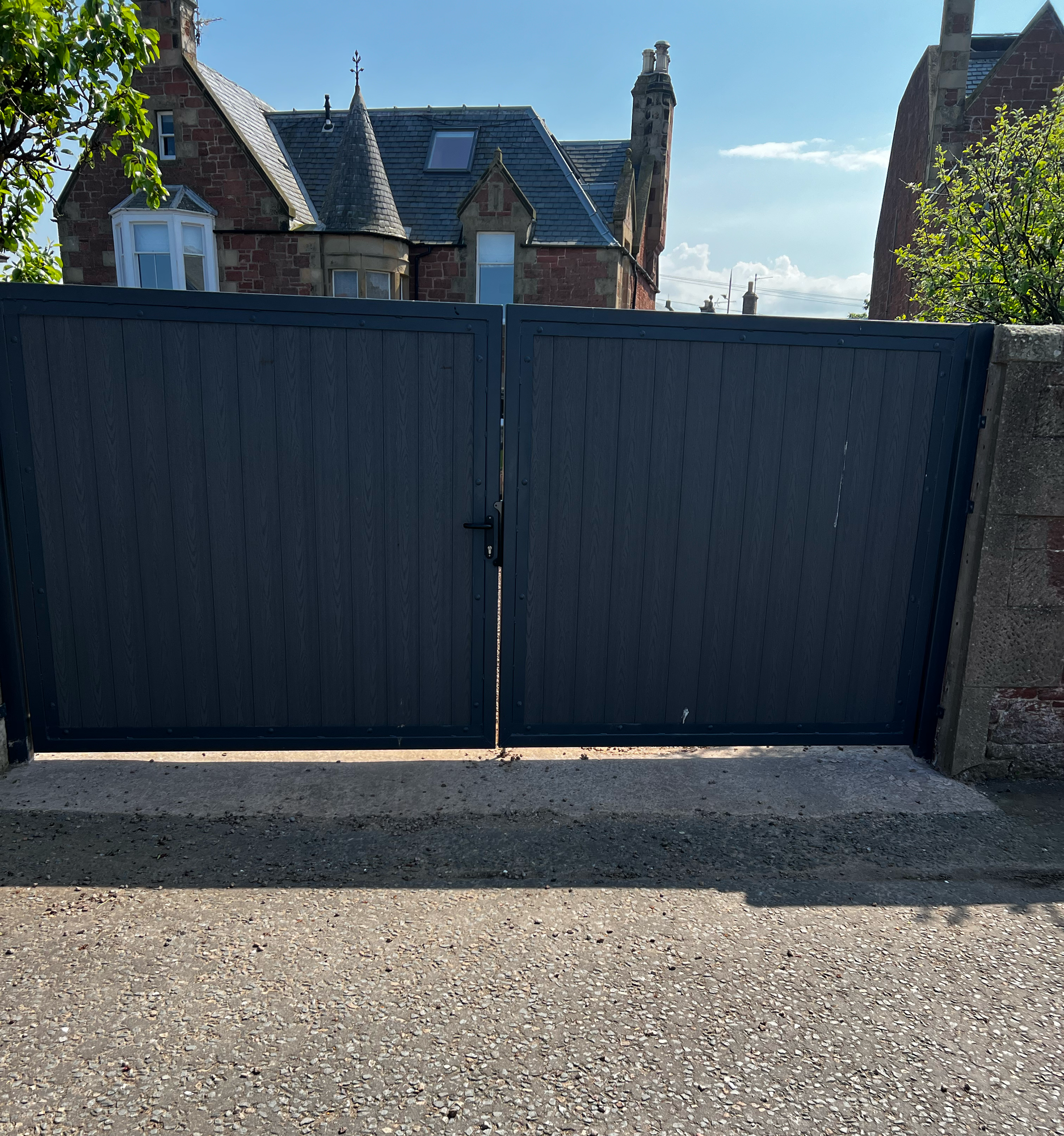 A black gate with a lock in front of a large brick house with a turret, dormer windows, and a slate roof. Green trees are visible on either side, and the sky is clear with some clouds.