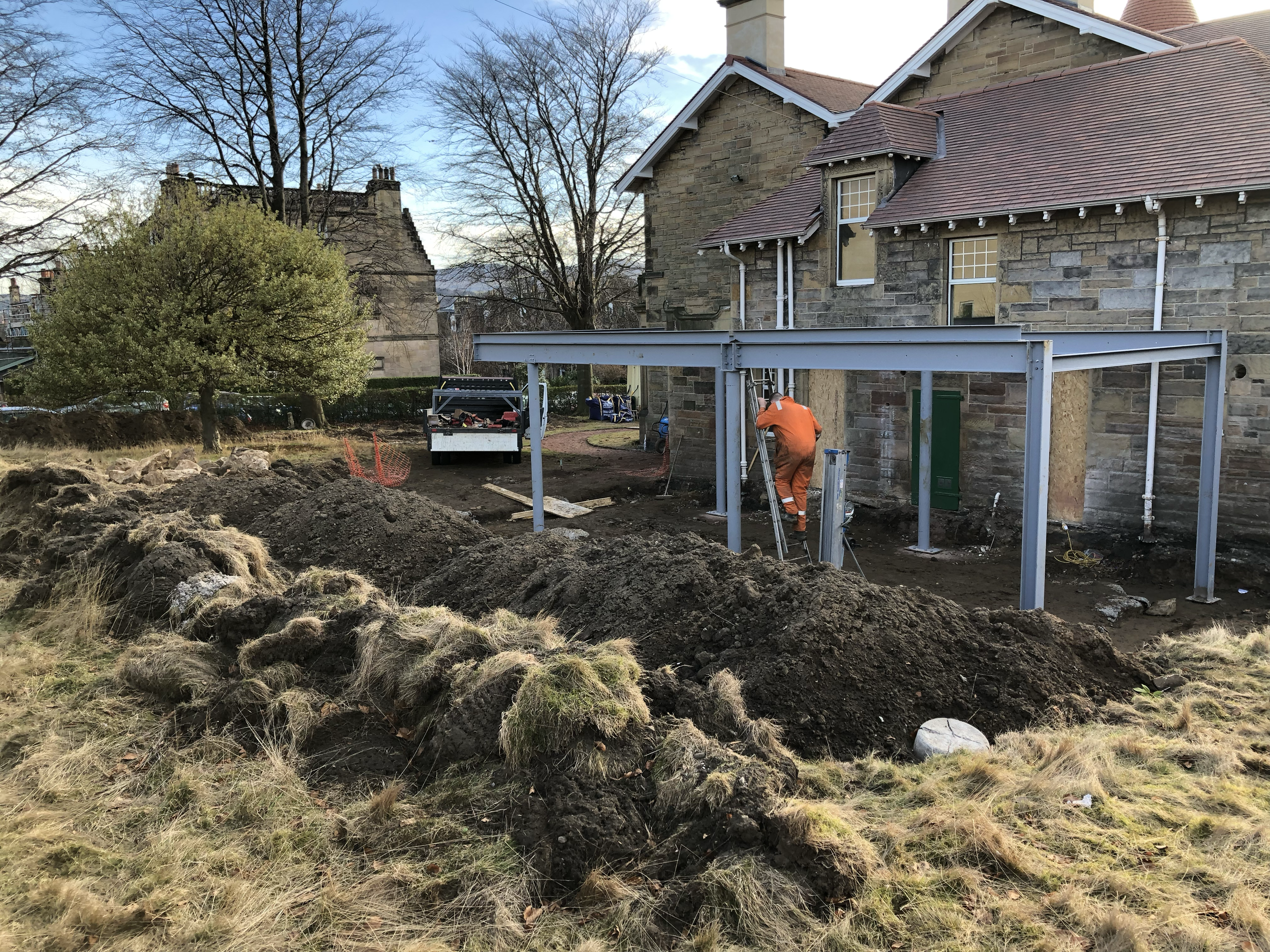 Construction work happening outside a stone house with a red-tiled roof, a worker in orange safety clothing, and an open area with disturbed soil and construction tools.
