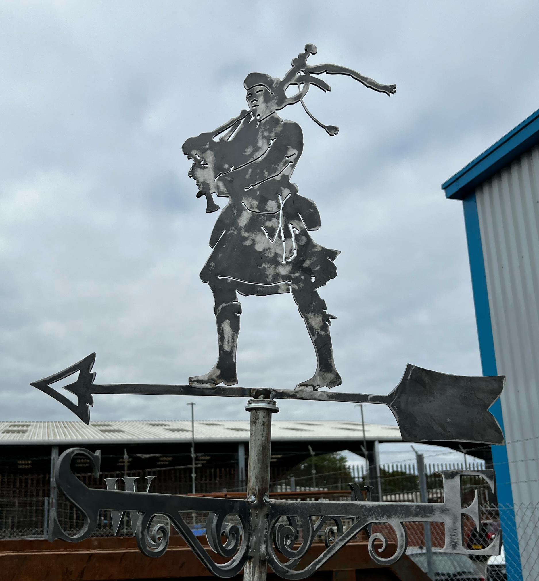 Metal weather vane depicting a girl with a hat holding a sword, mounted on a rooftop with clouds in the background.