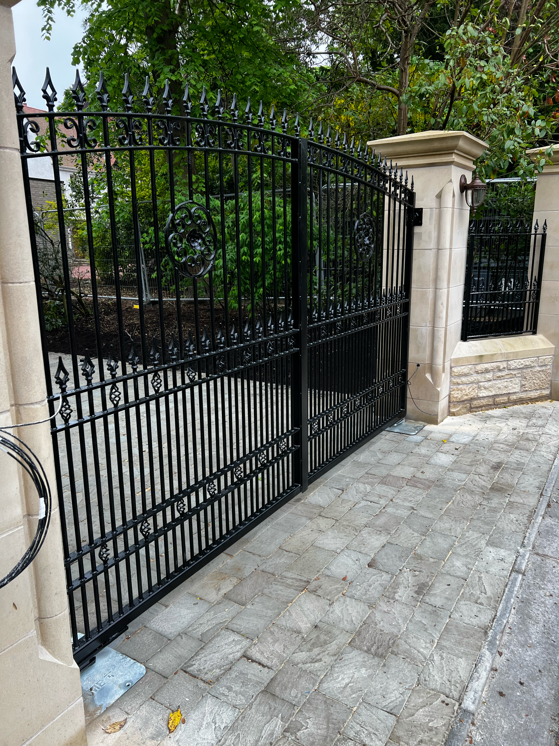 Black wrought iron gate with decorative circular and spearhead finials, supported by stone columns, with greenery and a tree in the background, and a cobblestone sidewalk in the foreground.