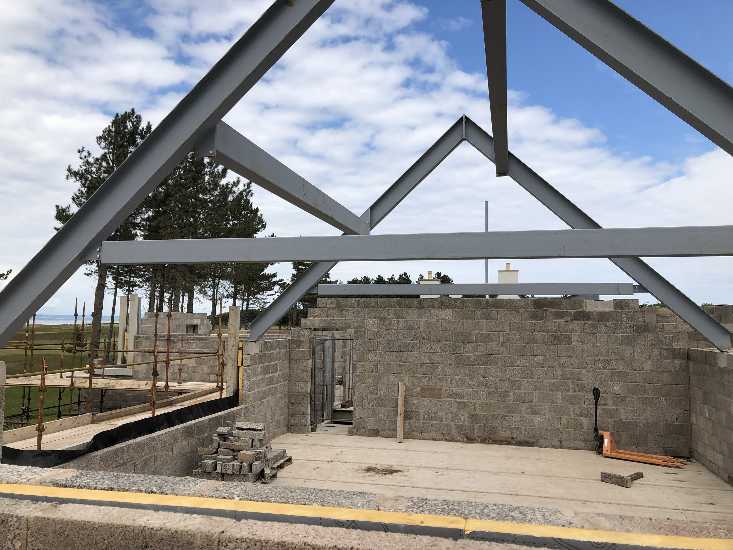 Construction site with brick walls and steel framework for a building, with trees and cloudy sky in the background.
