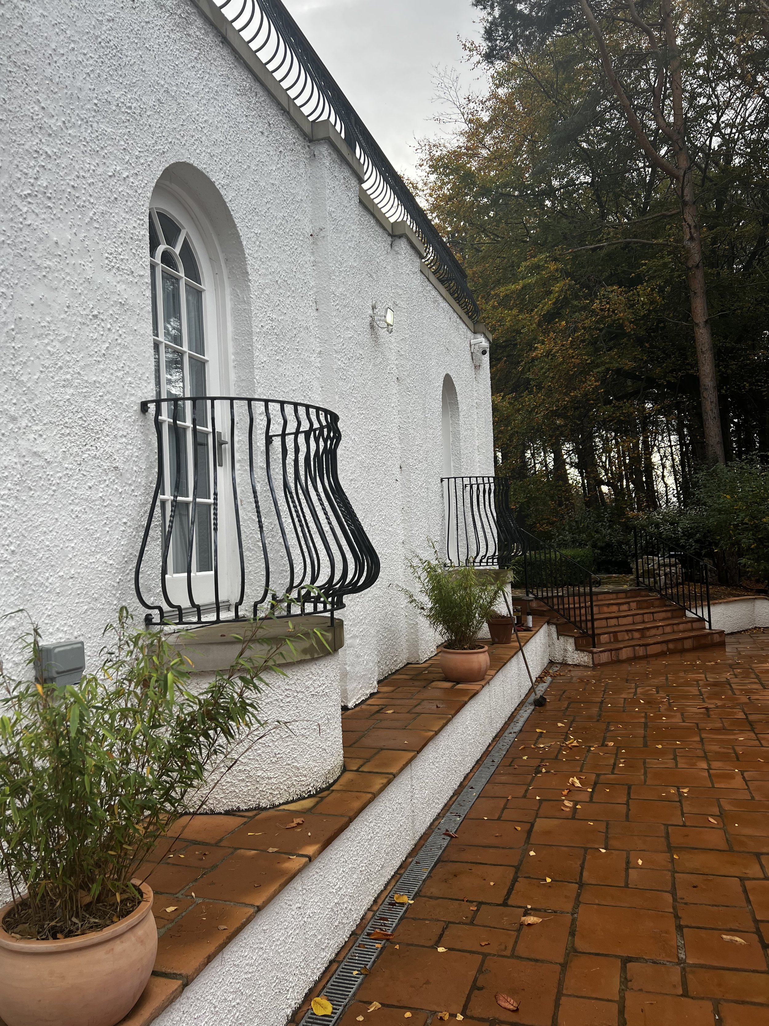Exterior view of a white stucco house with arched windows and small balconies with black iron railings. There is a brick pathway with fallen leaves, potted plants, and a drainage grate along the side of the house, set against a backdrop of trees with autumn foliage and a cloudy sky.
