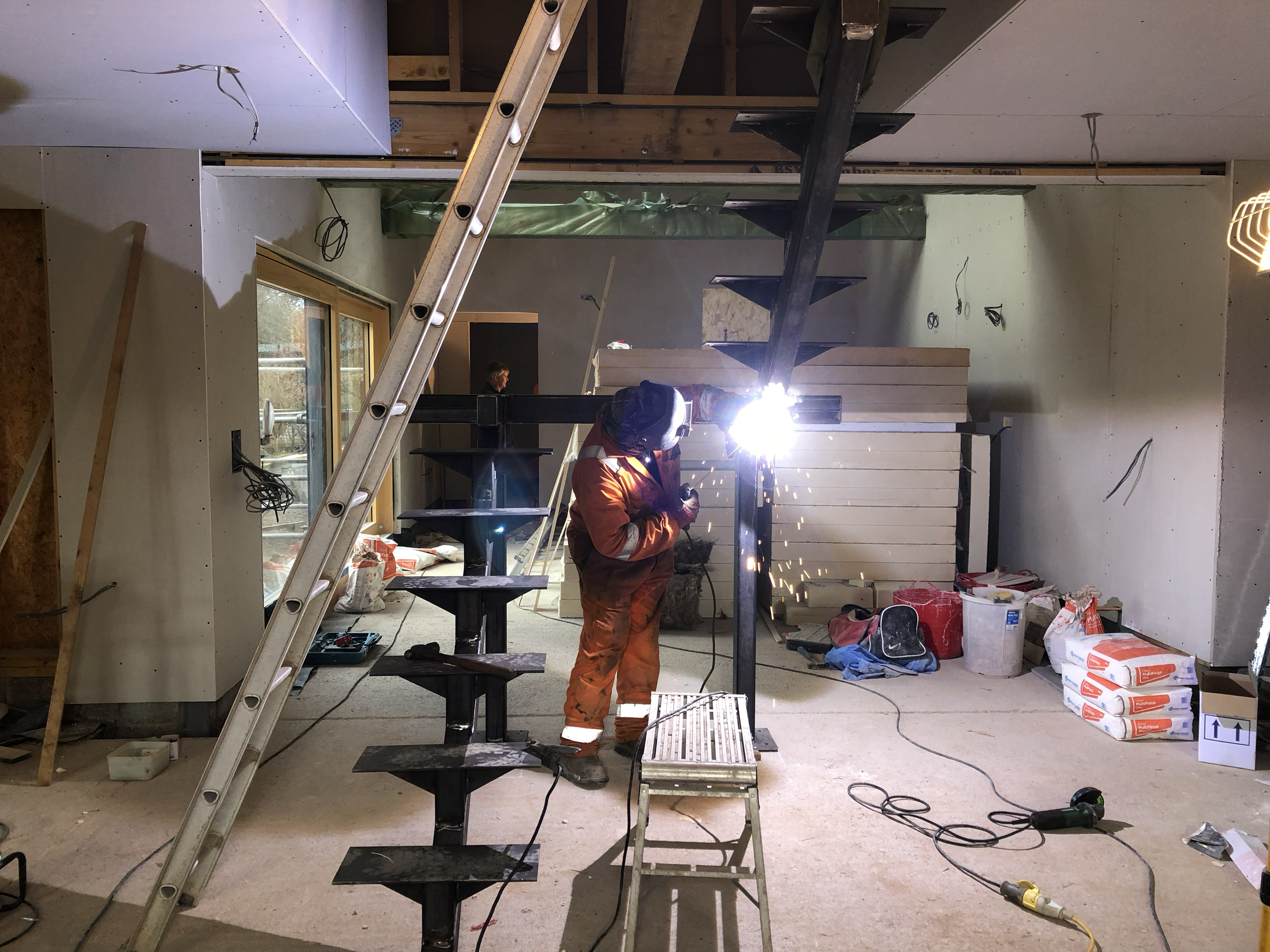Interior of a construction site with workers welding metal stairs, various construction materials, and tools scattered around.