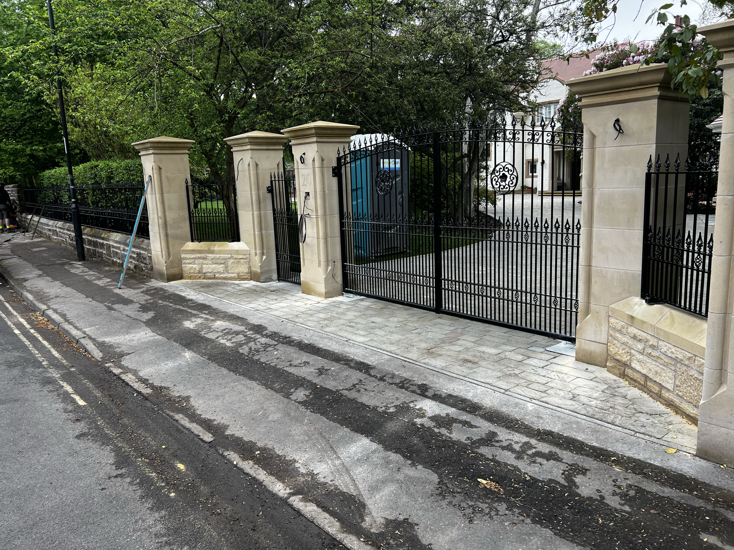 A gated entrance to a residential property with stone columns and black wrought iron gate, surrounded by trees and greenery, located next to a street with sidewalk and parked infrastructure.