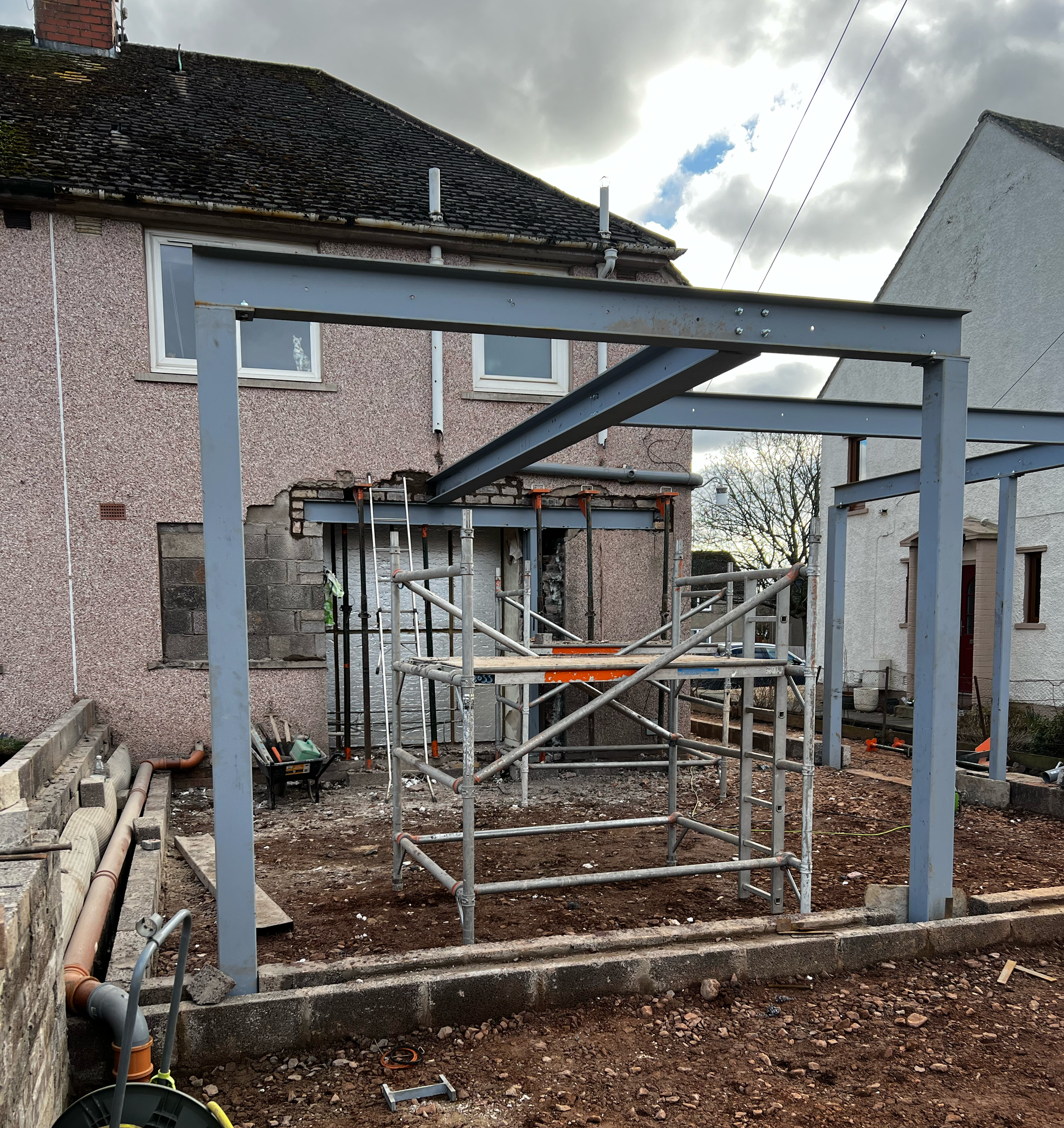 Construction site with metal framework and scaffolding in a backyard, adjacent to a residential building with a pebbled wall, two windows, and a newly dug foundation area.