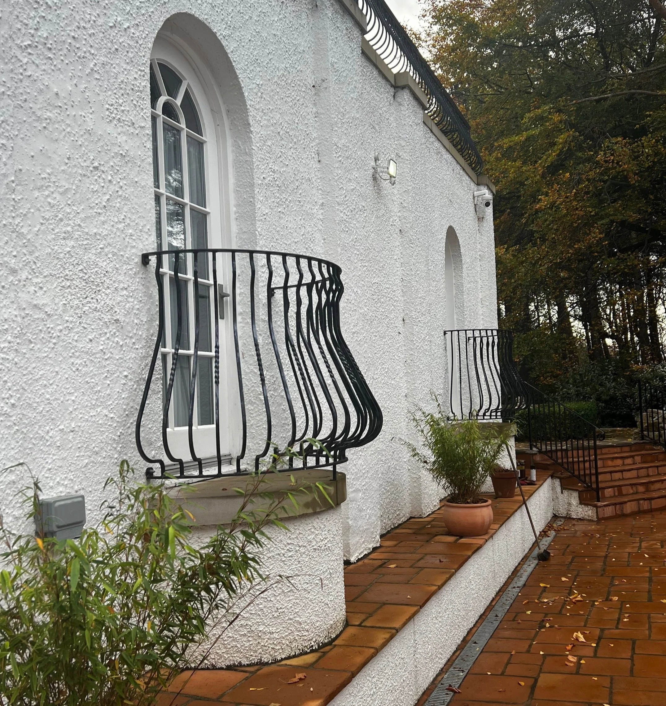 A white, textured exterior wall of a house with arched windows and small balconies with wrought iron railings. The house has a brick-paved pathway and some potted plants, with trees in the background that have green and orange leaves indicating fall.