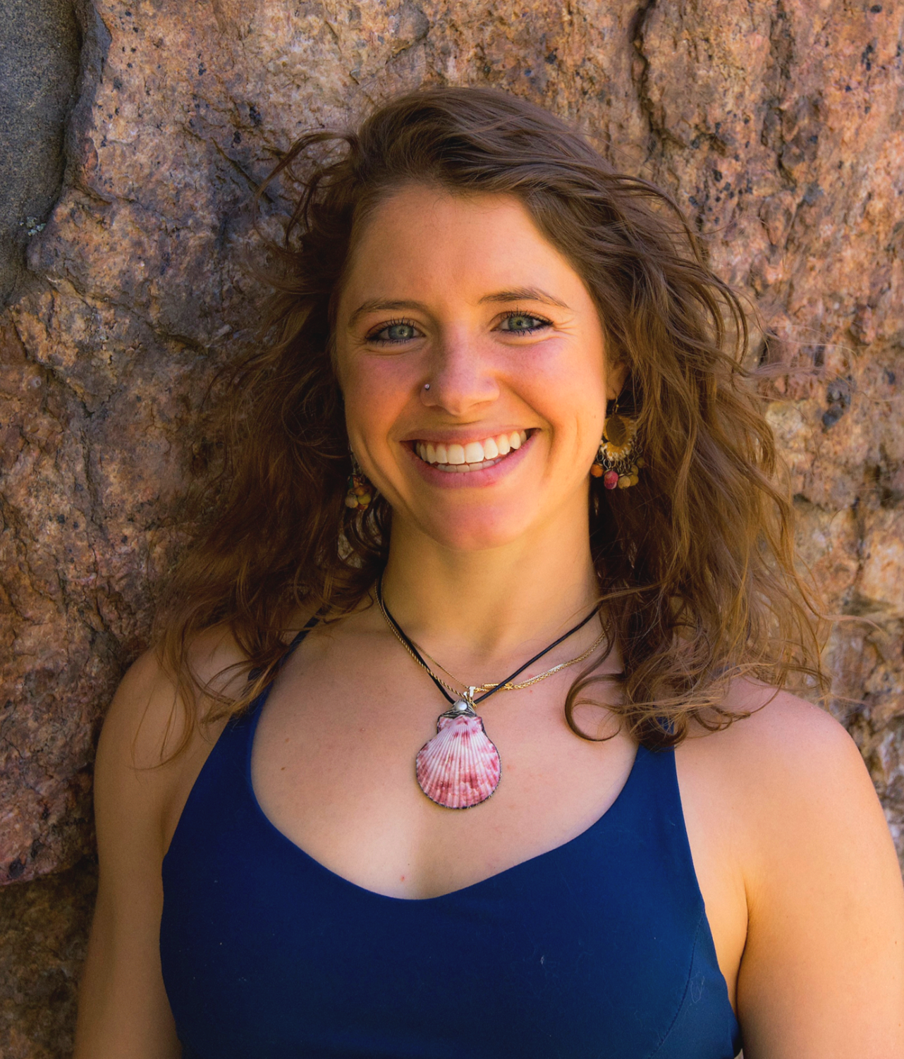 A woman with curly brown hair and blue eyes, smiling and leaning against a rocky background, wearing a blue tank top and a shell necklace.