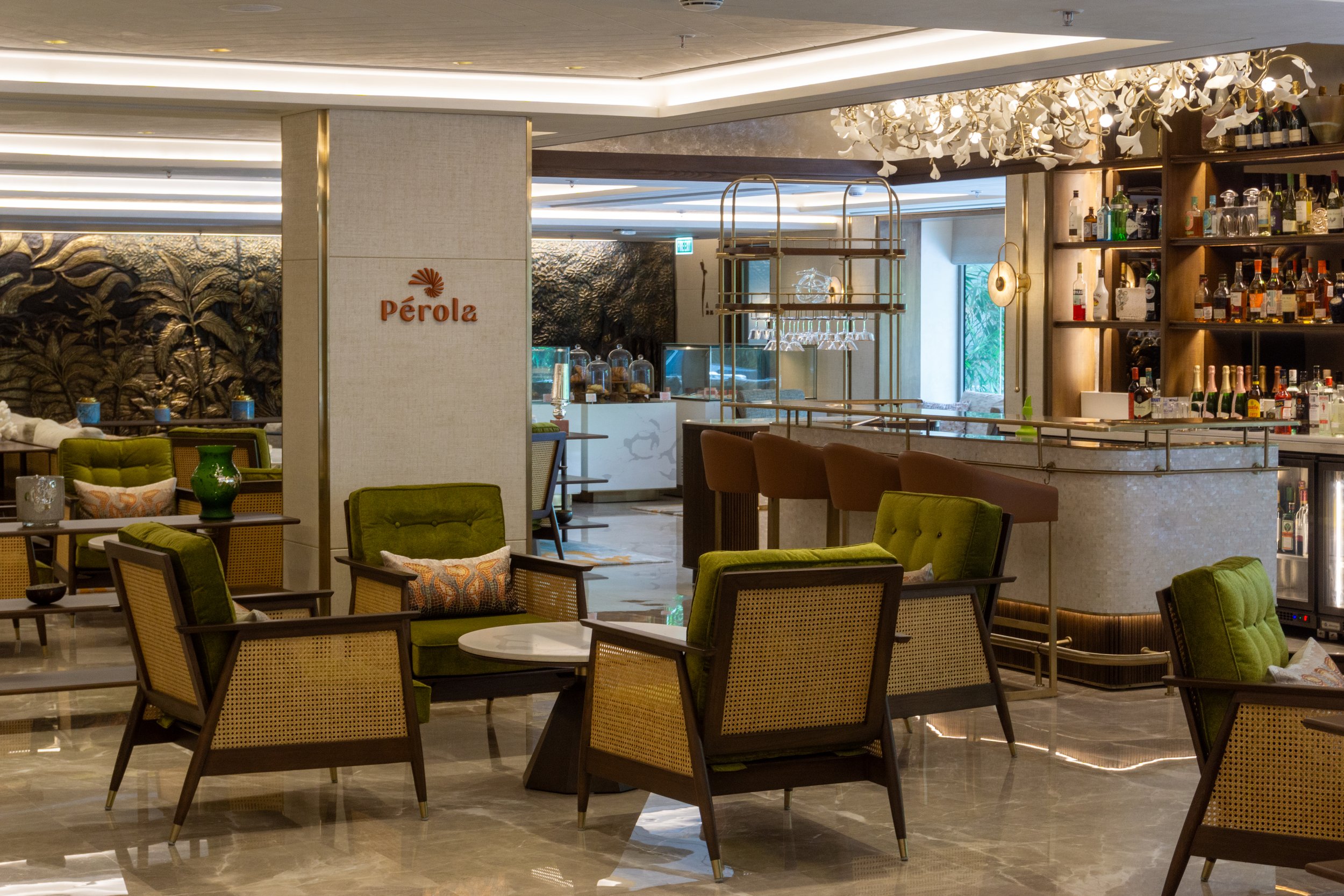 Interior of a stylish bar and lounge area with green and beige chairs, decorative pillows, a bar with various bottles, and a sign that reads 'Pérola'.