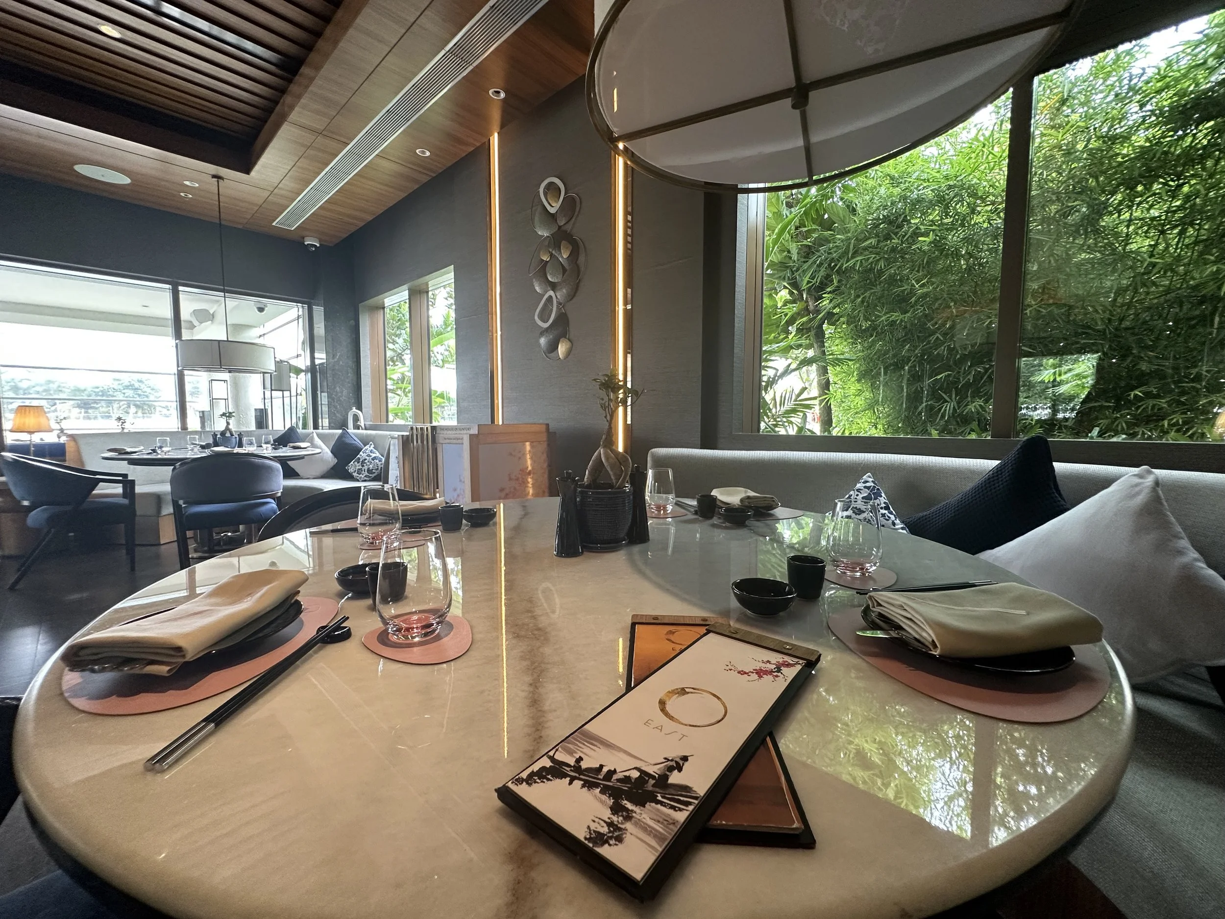 Upscale restaurant table set with glasses, napkins, chopsticks, and menus, featuring large windows with greenery outside and modern interior decor.