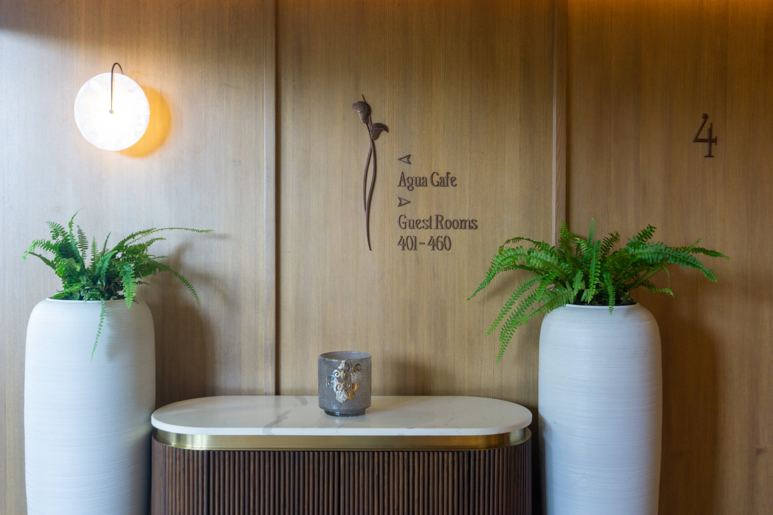 Interior hotel lobby with wooden wall, two large white vases with green ferns, a marble-topped side table, wall-mounted sign with directions to Agua Cafe and guest rooms 401-460, and a modern wall sconce.