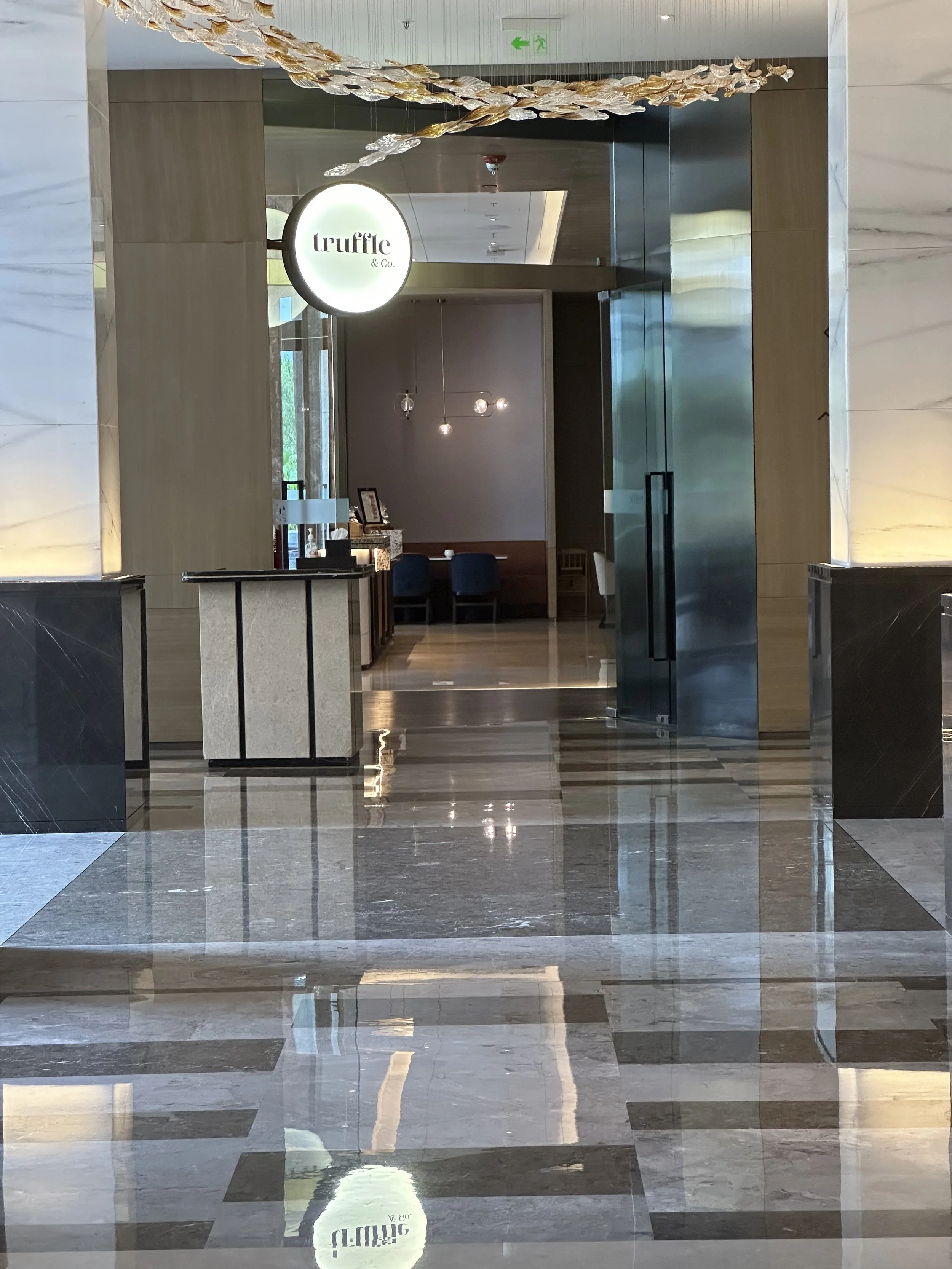 Interior view of a modern restaurant or lounge area with a chandelier overhead and a sign reading "truffle & Co." in the background, with polished marble floors and wooden accents.
