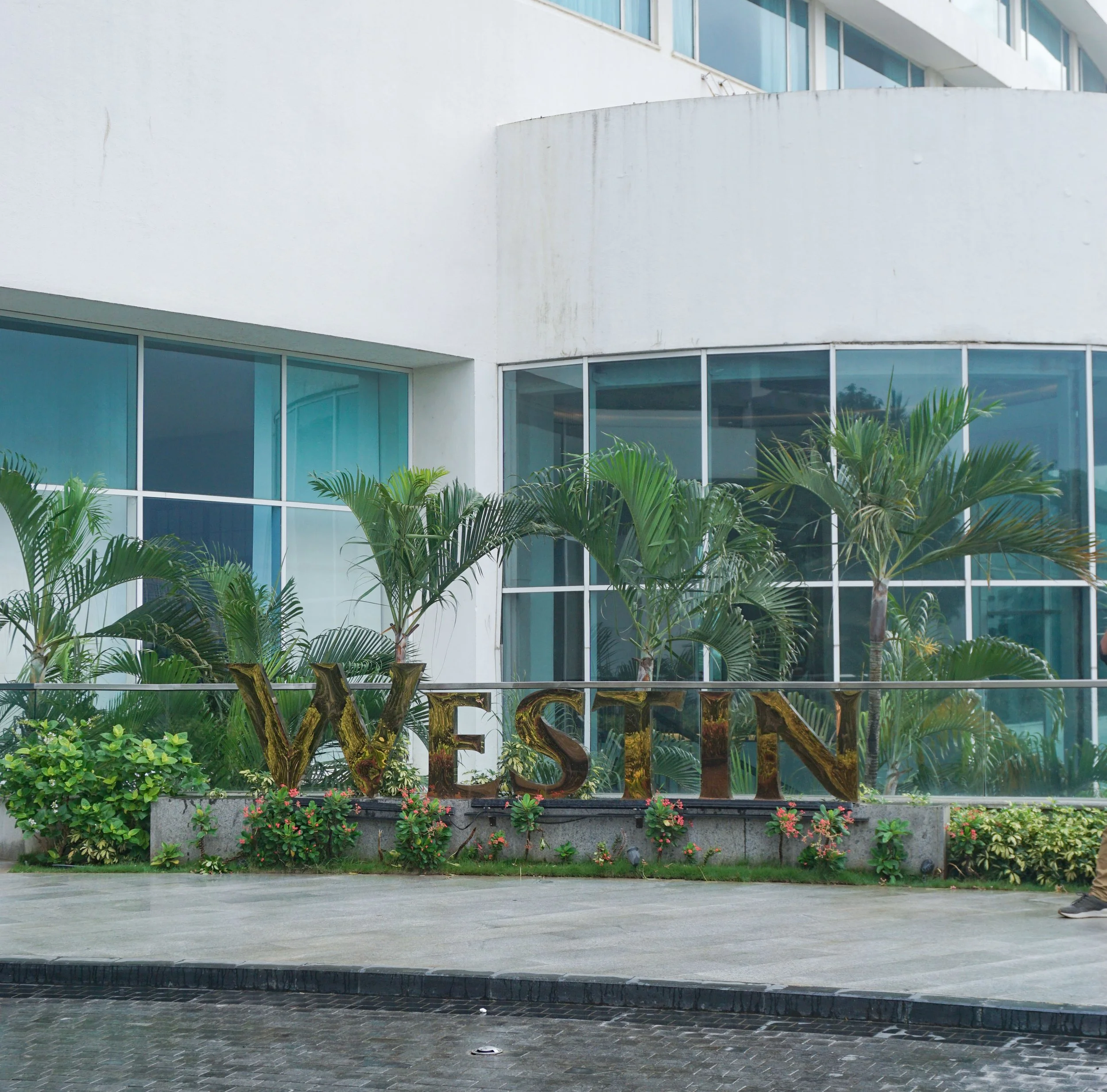 Exterior of a modern building with large glass windows, tropical plants, and a decorative sign spelling 'WESTIN' in gold-colored letters.