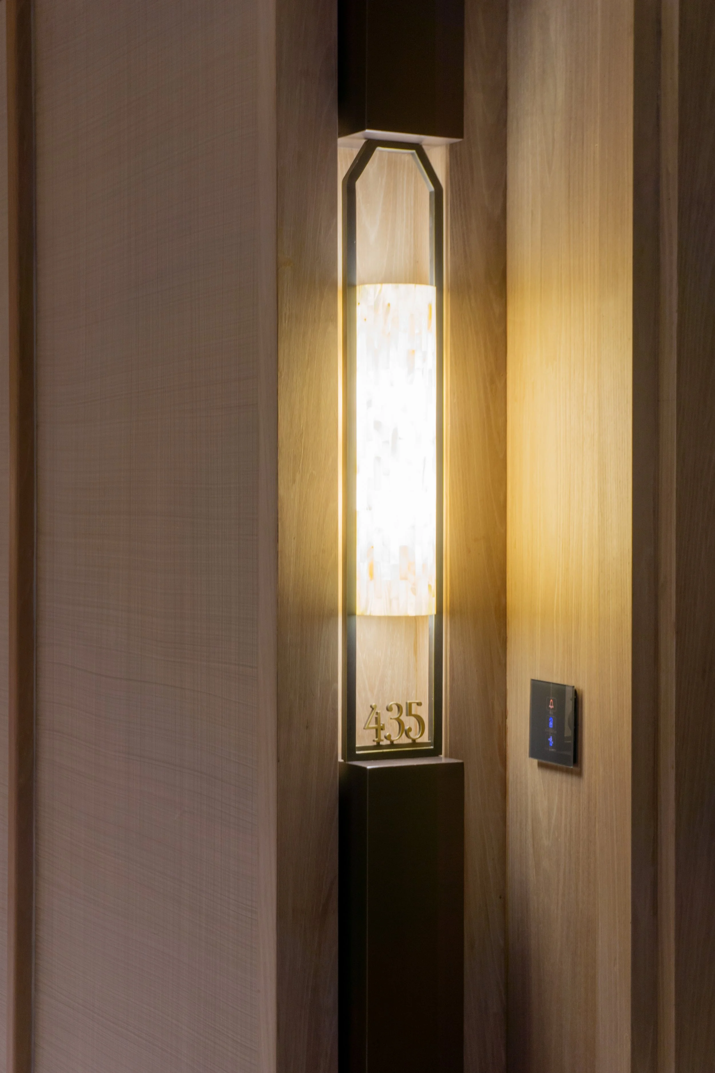A modern wall-mounted lamp with a black frame around a lit, rectangular, decorative shade next to a digital elevator control panel, in a wood-paneled hallway with the number 435 displayed at the base of the lamp.