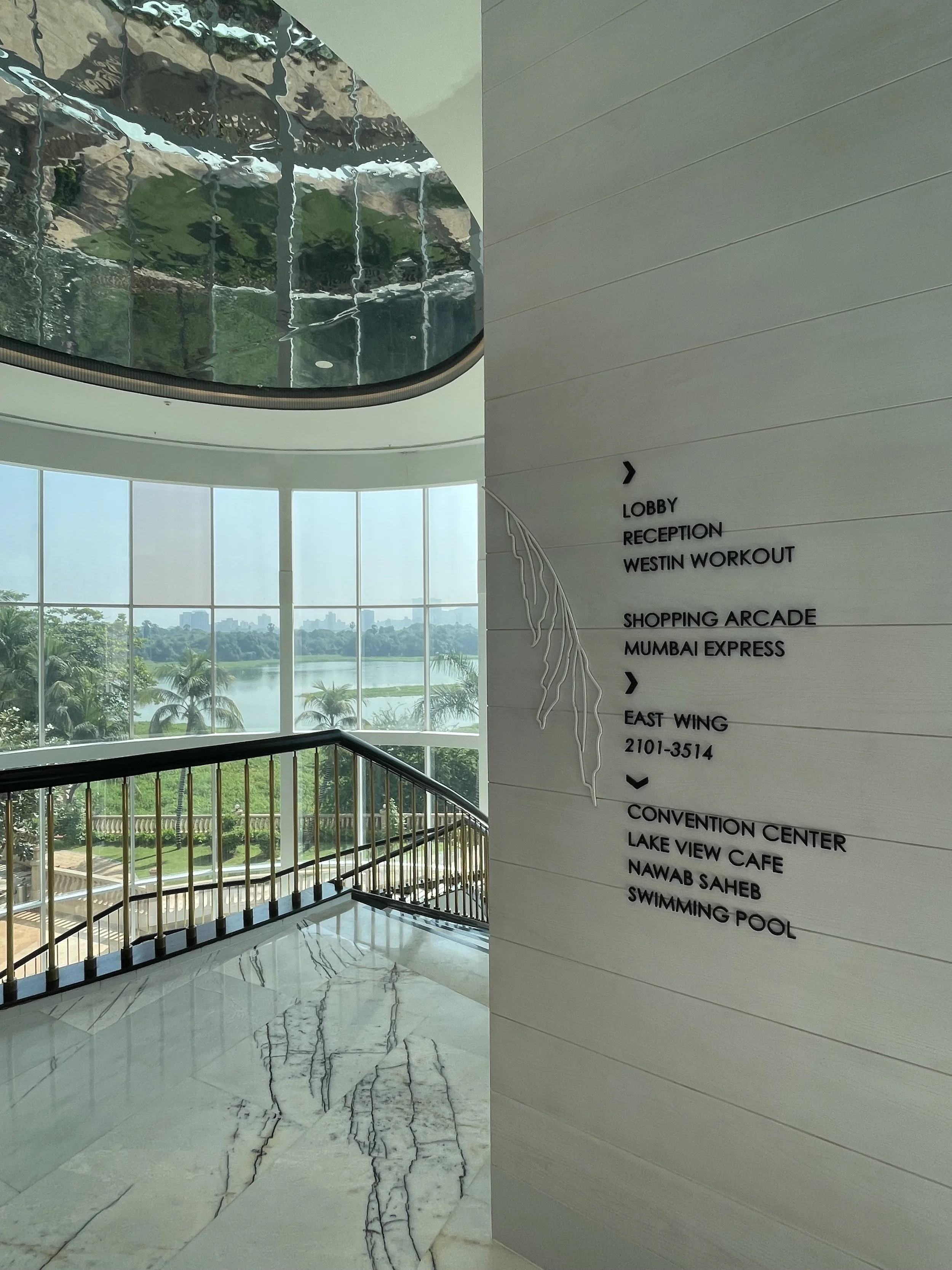 Indoor space with a staircase, large window view of trees and river outside, and a directory wall with black text indicating directions to various amenities and areas inside a building.