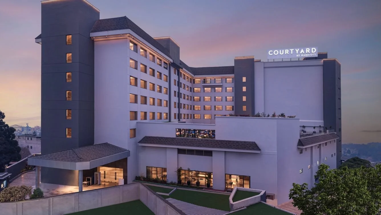 The exterior of a Marriott Courtyard hotel building during sunset, with a sign illuminated on the rooftop. The building has multiple floors with many windows and a well-lit entrance area.