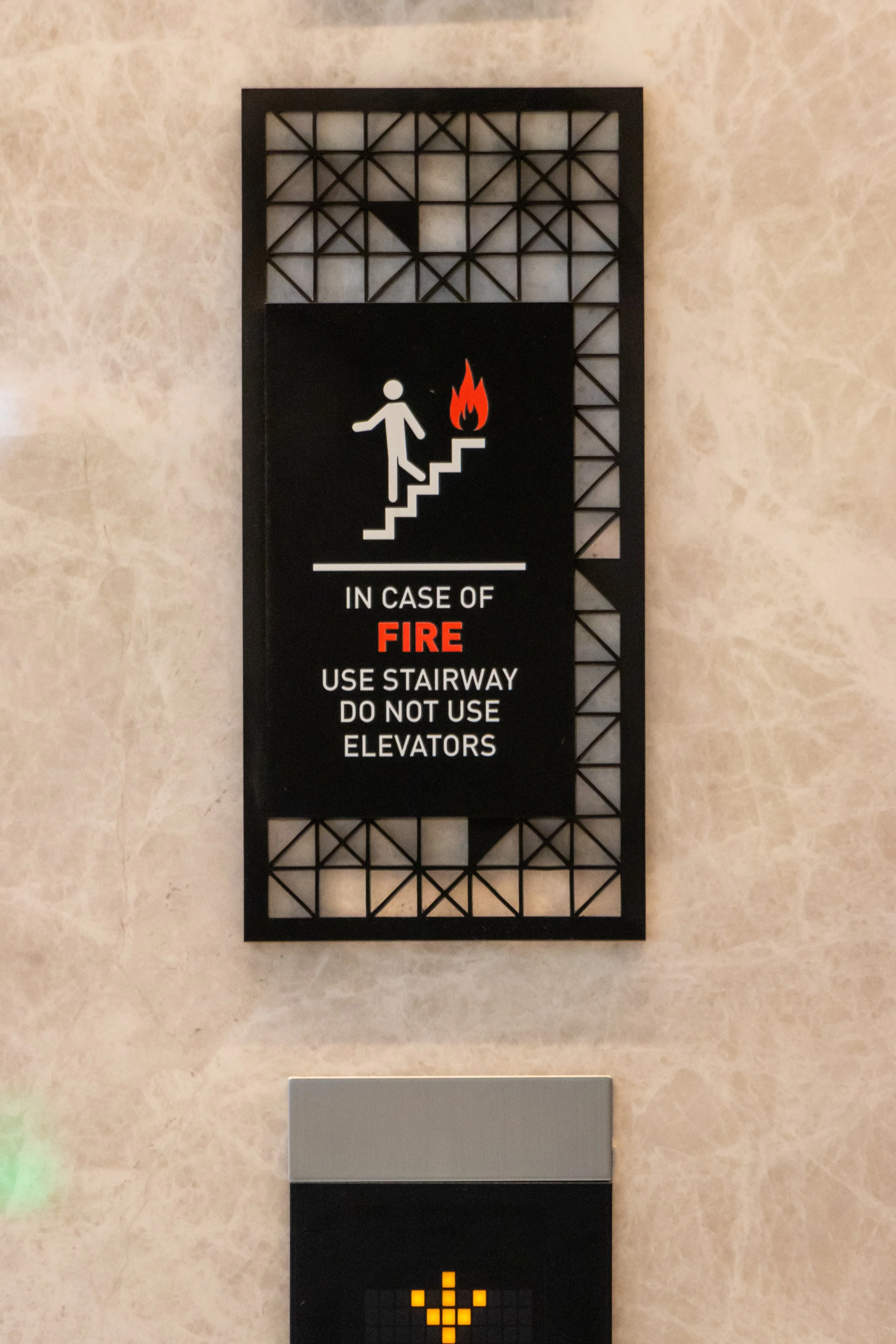 Fire safety sign with a person going up stairs and a flame, indicating in case of fire use stairs, do not use elevators, mounted on a beige wall.