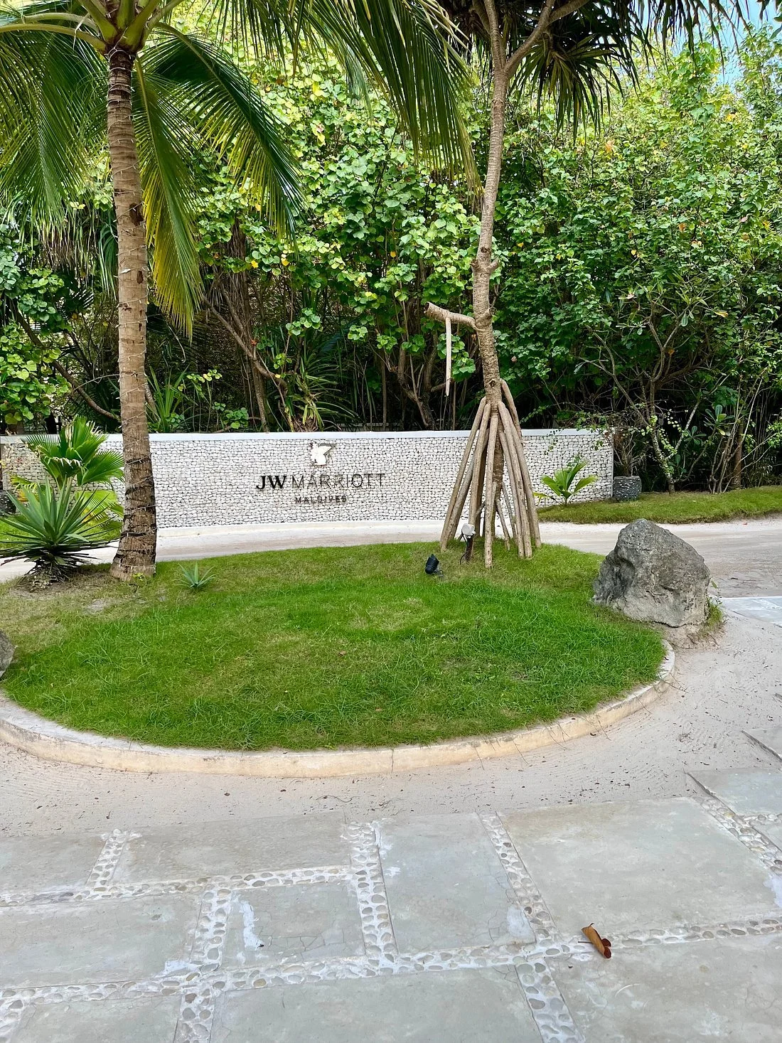 “JW Marriott Maldives” sign on a white stone wall surrounded by palm trees and tropical plants, with a small circular grassy area and large rock in front of a sidewalk.