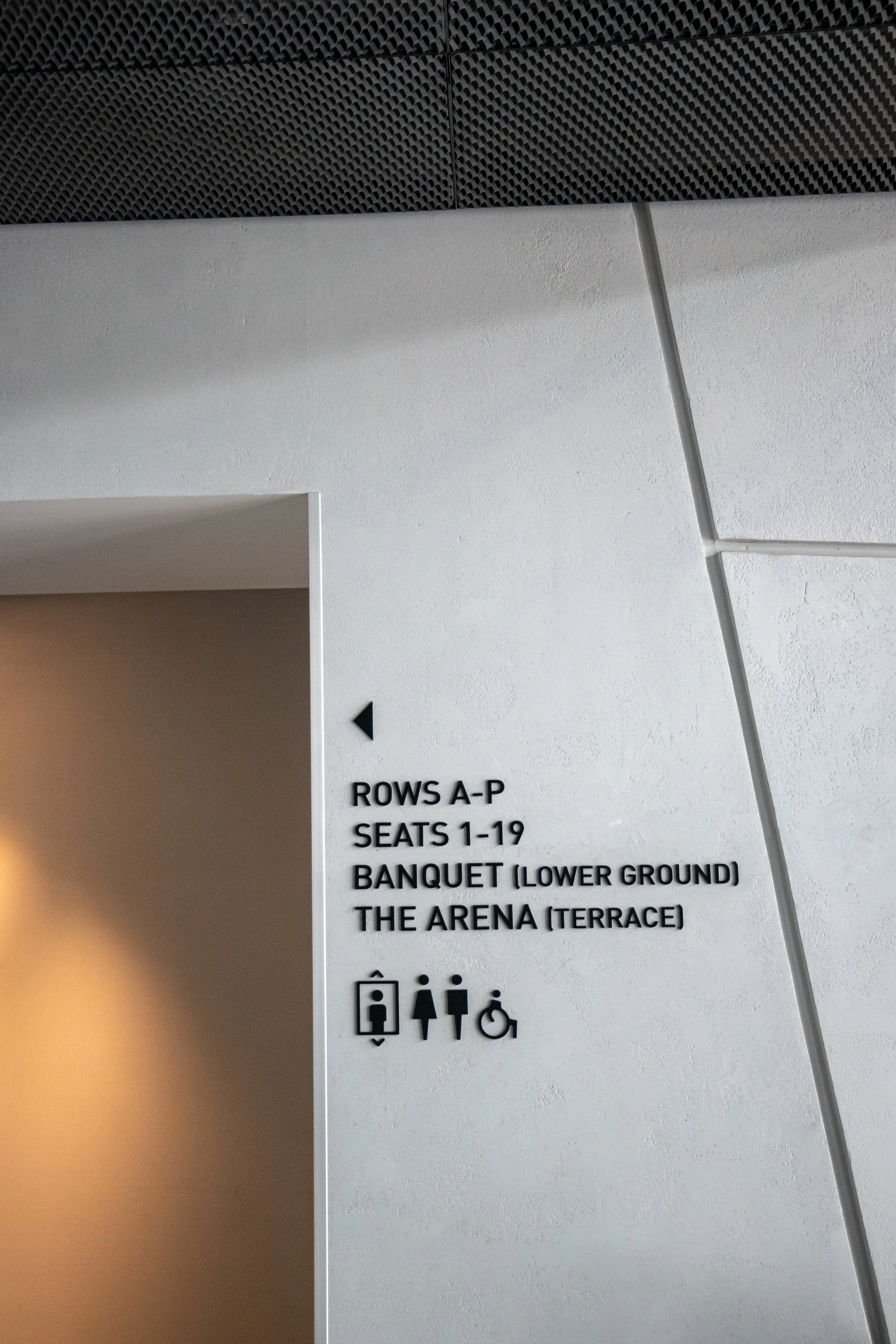Sign indicating directions to rows A-P, seats 1-19, banquet lower ground, and the arena terrace, with icons for elevator, women, men, and wheelchair accessible restroom.