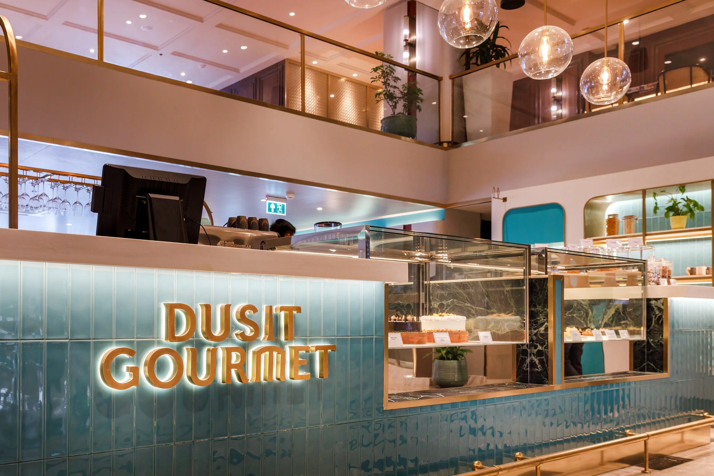 Inside a bakery called Dusit Gourmet with cakes on display behind glass counters and a modern decor with hanging glass light fixtures.