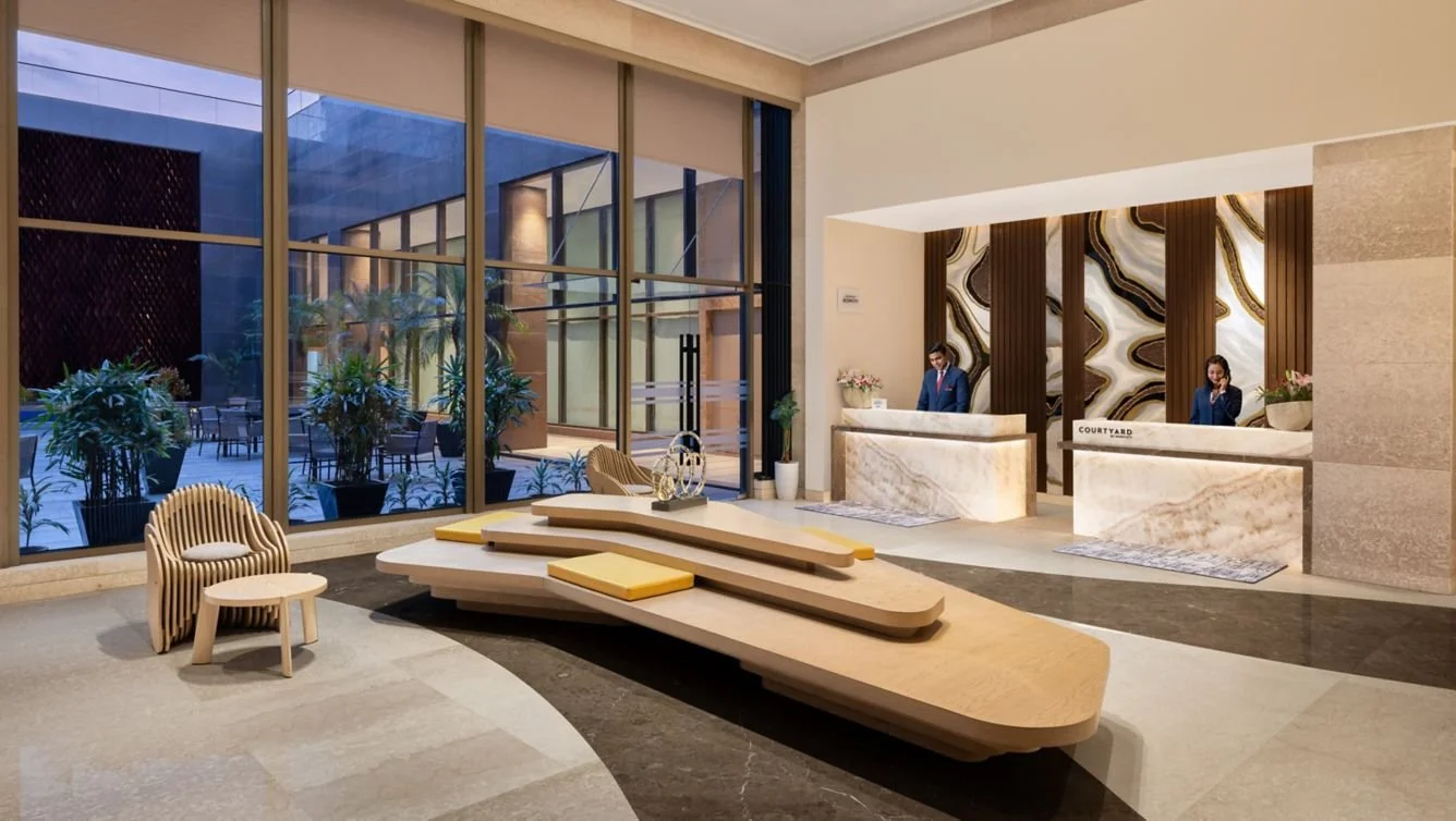 Hotel lobby with reception desk, two staff members, large windows, and modern decor.
