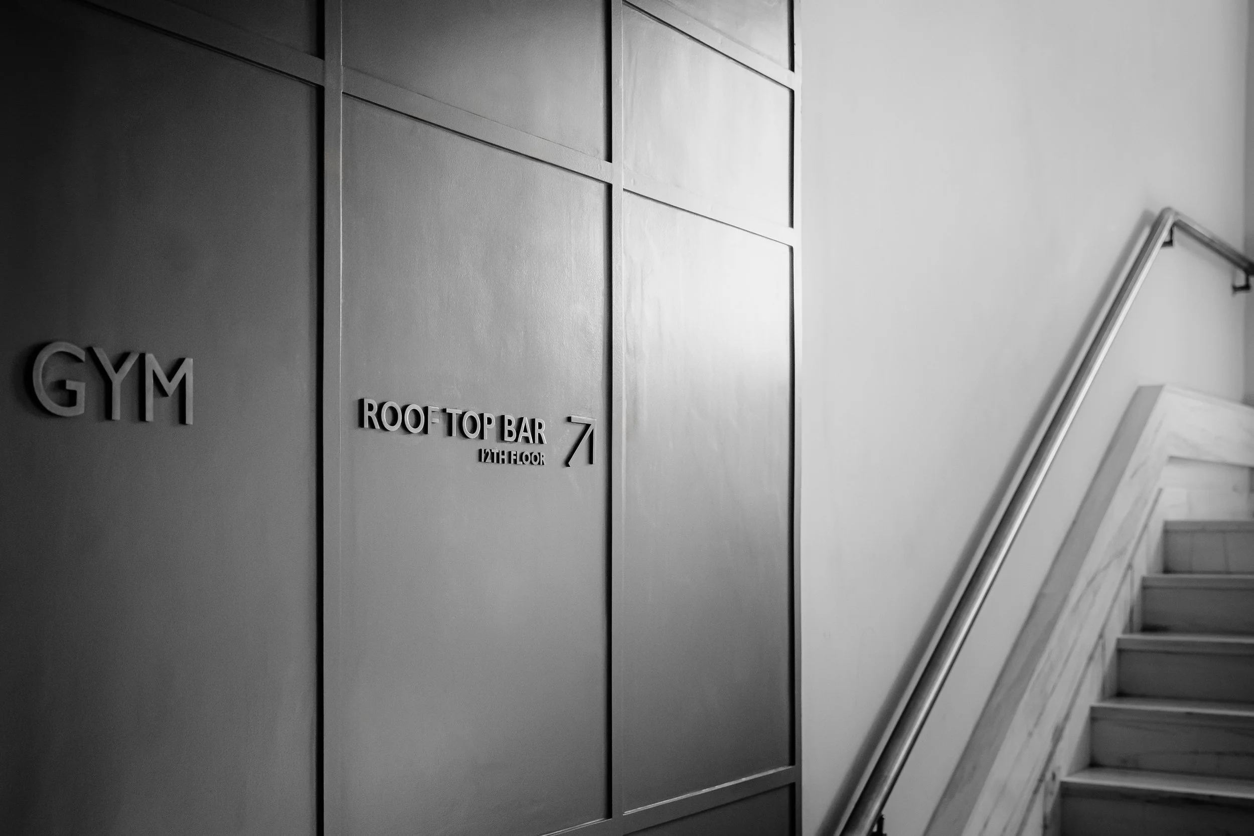 Black and white photo of a staircase next to a wall with signage indicating a gym and rooftop bar on the 12th floor, pointing to the right.