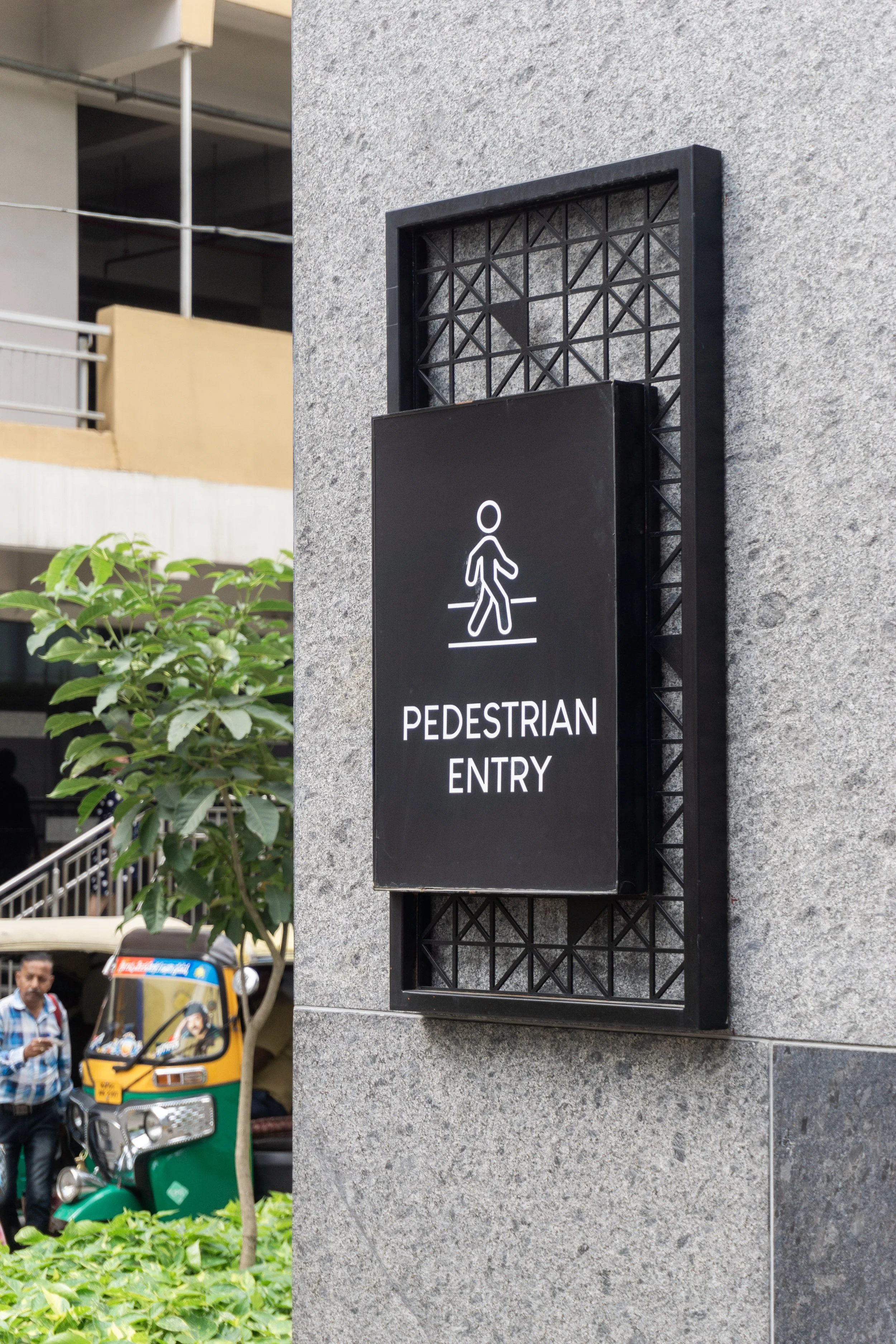 A black sign that reads 'PEDESTRIAN ENTRY' with a walking person icon, mounted on a grey stone wall outside.