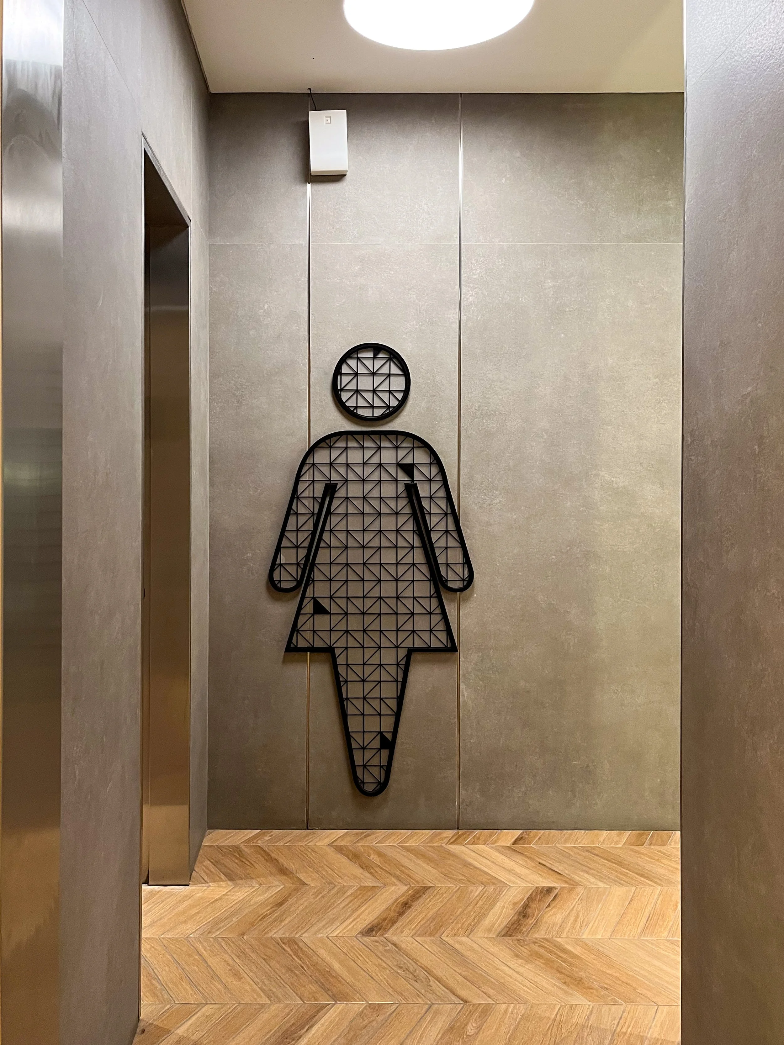 Decorative wall art of a female figure with a circle head, in an outline of a dress and arms, made of black geometric lines, displayed on a gray wall in an elevator lobby.