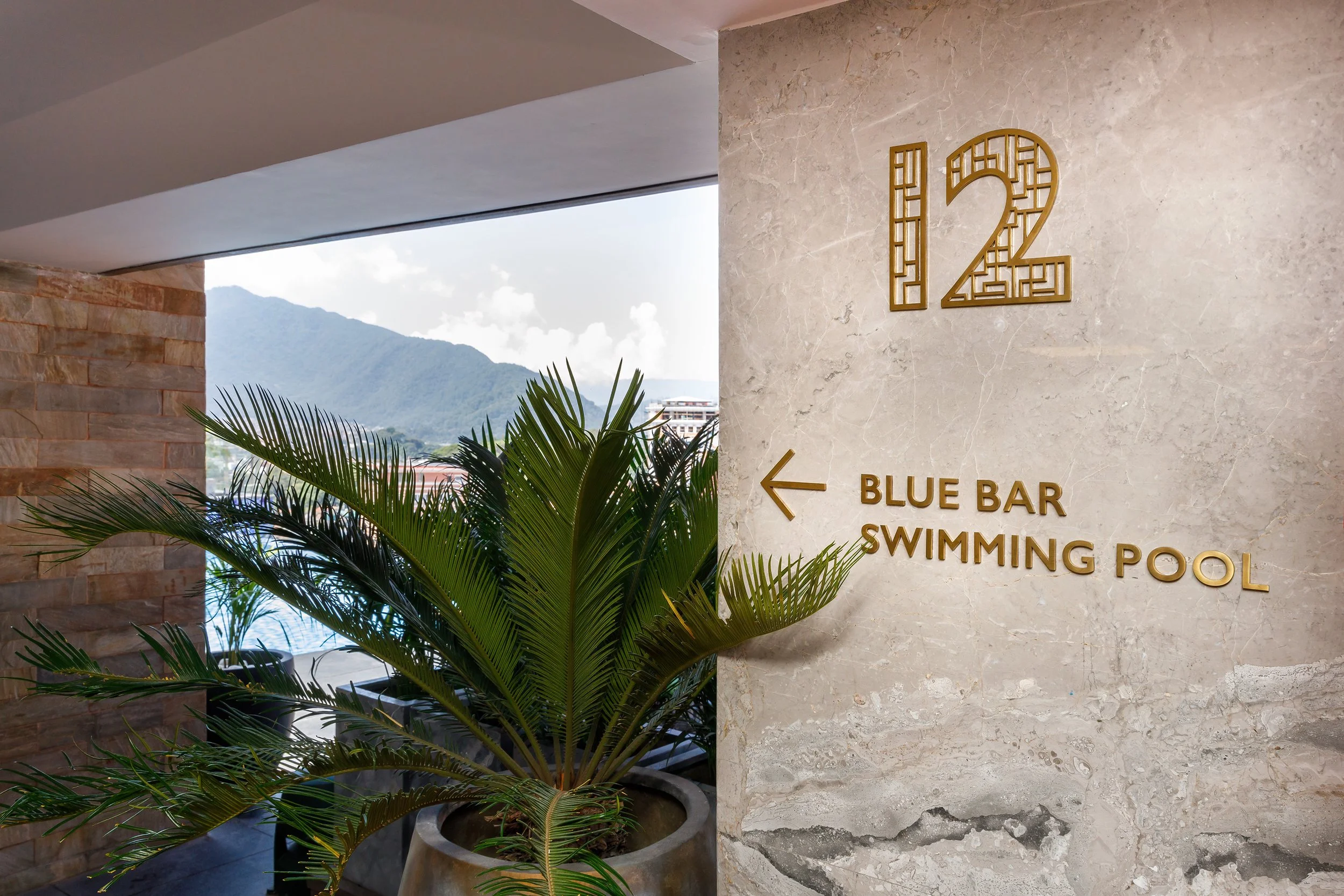 Sign on a beige marble wall with gold numbers '12' and gold text indicating directions to the 'Blue Bar' and 'Swimming Pool' with an arrow pointing left. A green potted palm plant is in front of the wall, and mountains are visible through a large win