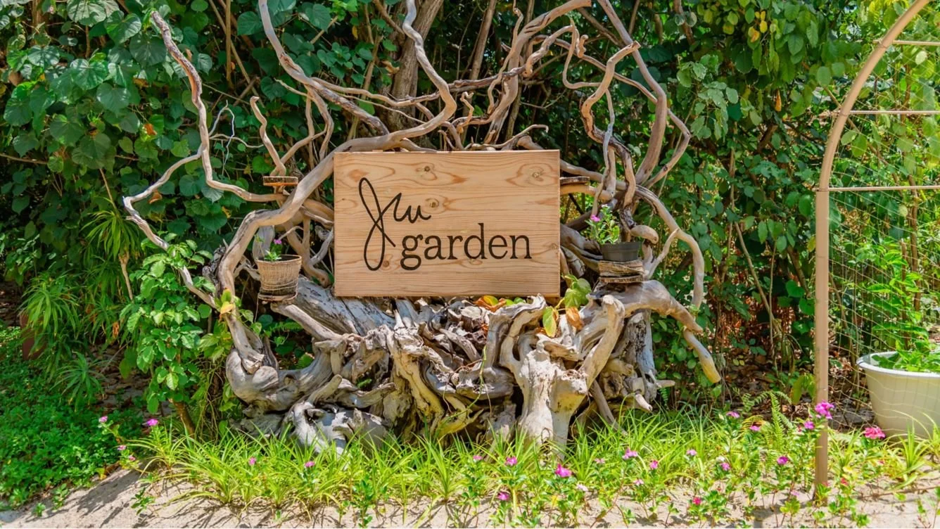 Wooden garden sign with the words "Jiu garden" attached to a large piece of driftwood or root surrounded by green plants, flowers, and garden tools.