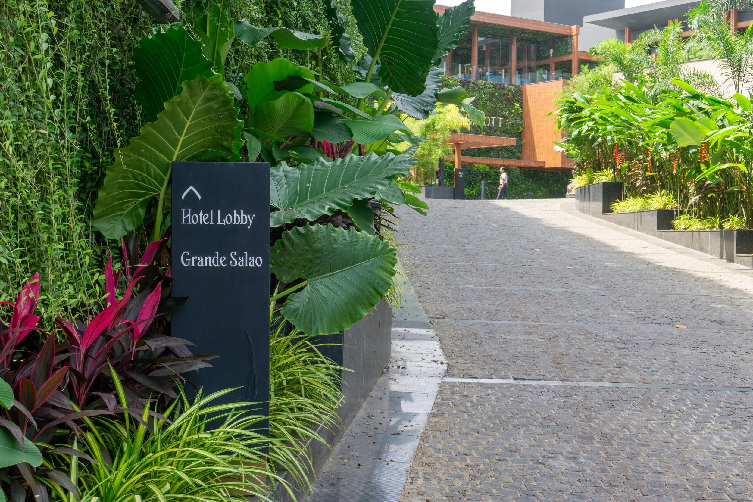 Hotel lobby sign next to lush green plants along a curved cobblestone walkway leading to a modern building in the background.