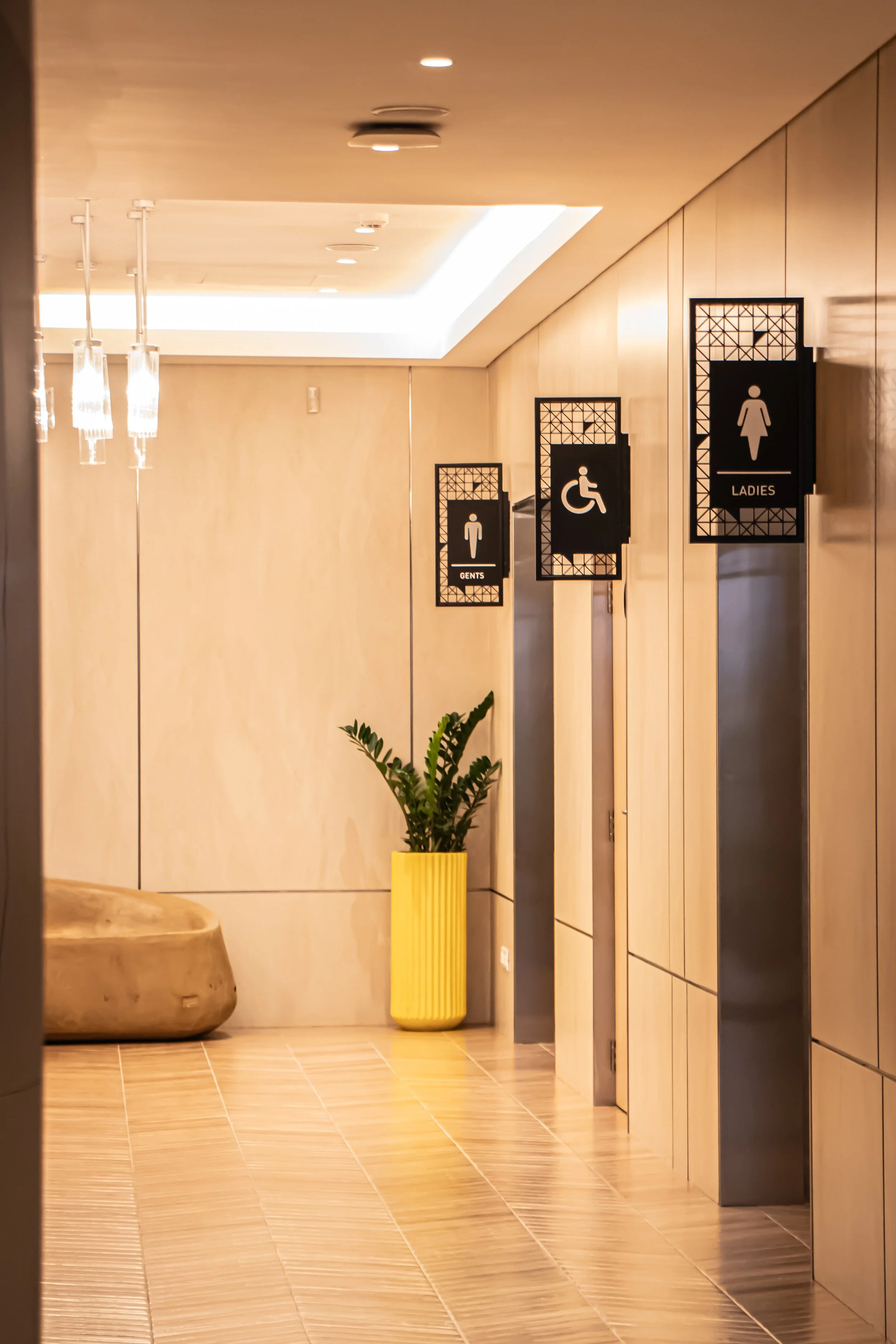 Public restroom hallway with signs for gents, ladies, and accessible toilets, and a yellow planter with a green plant.