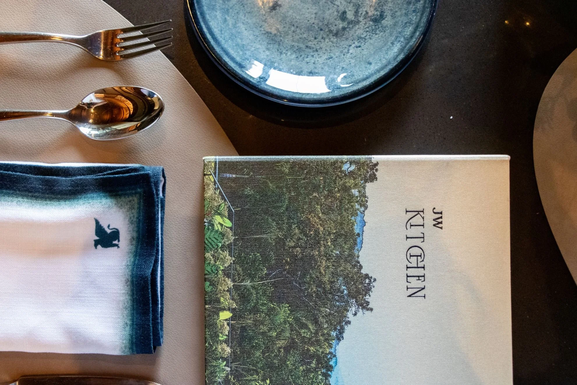Place setting with a menu, a napkin with a whale silhouette, a fork, and a spoon on a beige tablecloth, with a stone plate and a black plate in the background.