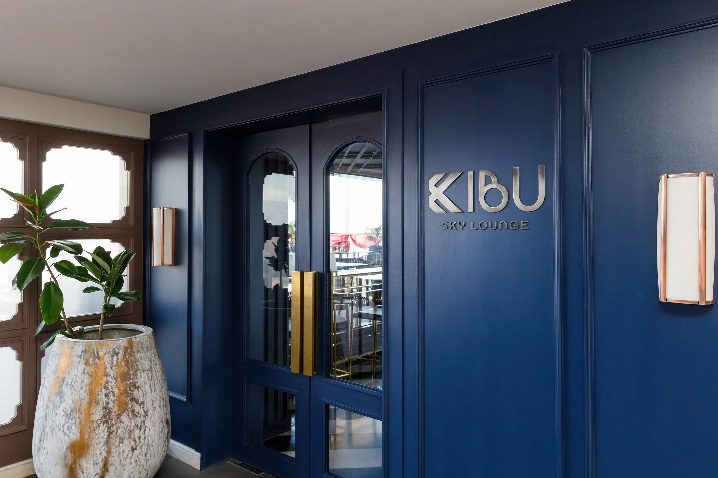 Entrance to Kibu Sky Lounge with blue walls, glass doors with gold handles, decorative wall sconces, and a large planter with a green plant.
