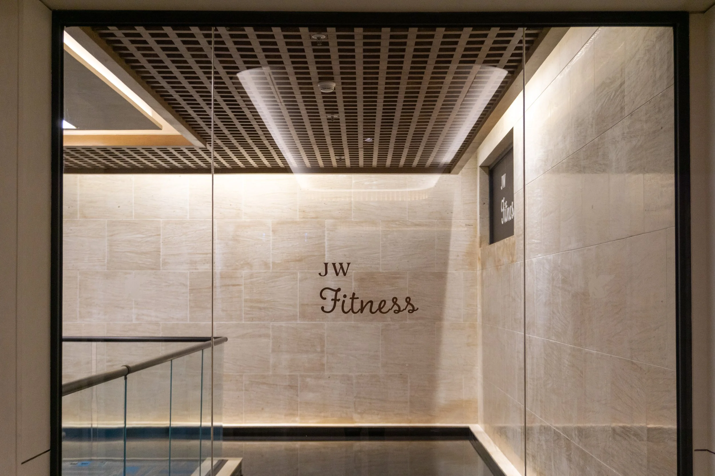 Glass window door with JW Fitness logo on beige tiled wall behind it, modern interior with wooden ceiling and glass railing.